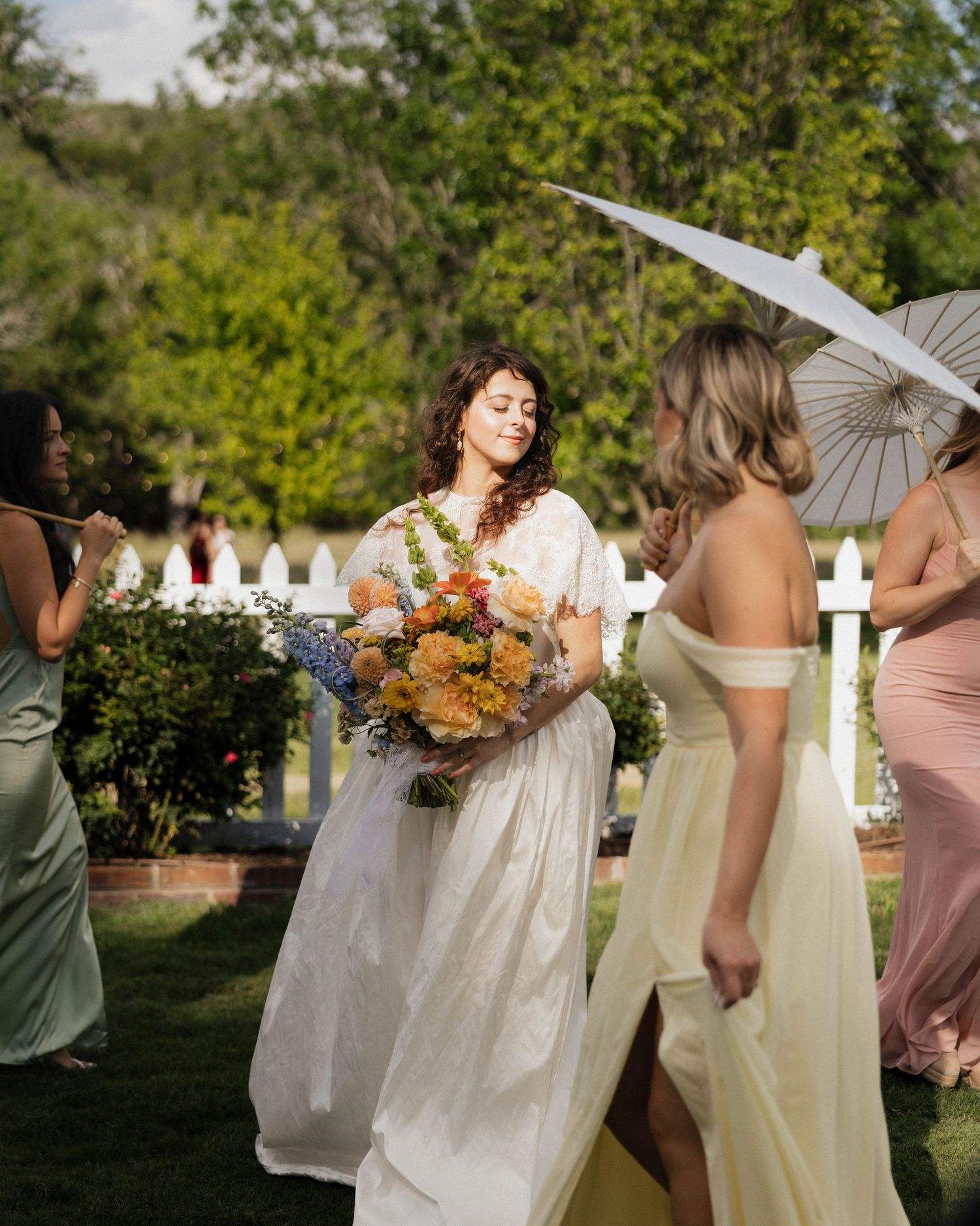 Bride in white gown holds colorful bouquet surrounded by bridesmaids in pastel dresses at outdoor garden wedding.
