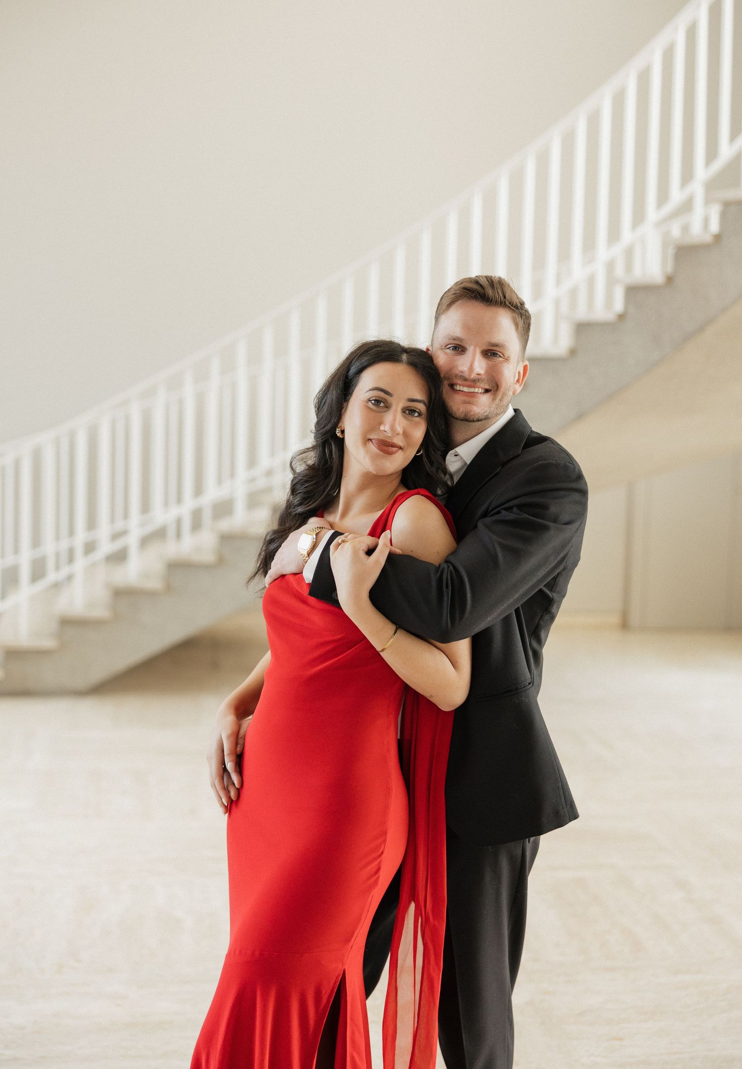 A couple in formal attire embraces in front of white stairs in an elegant setting.