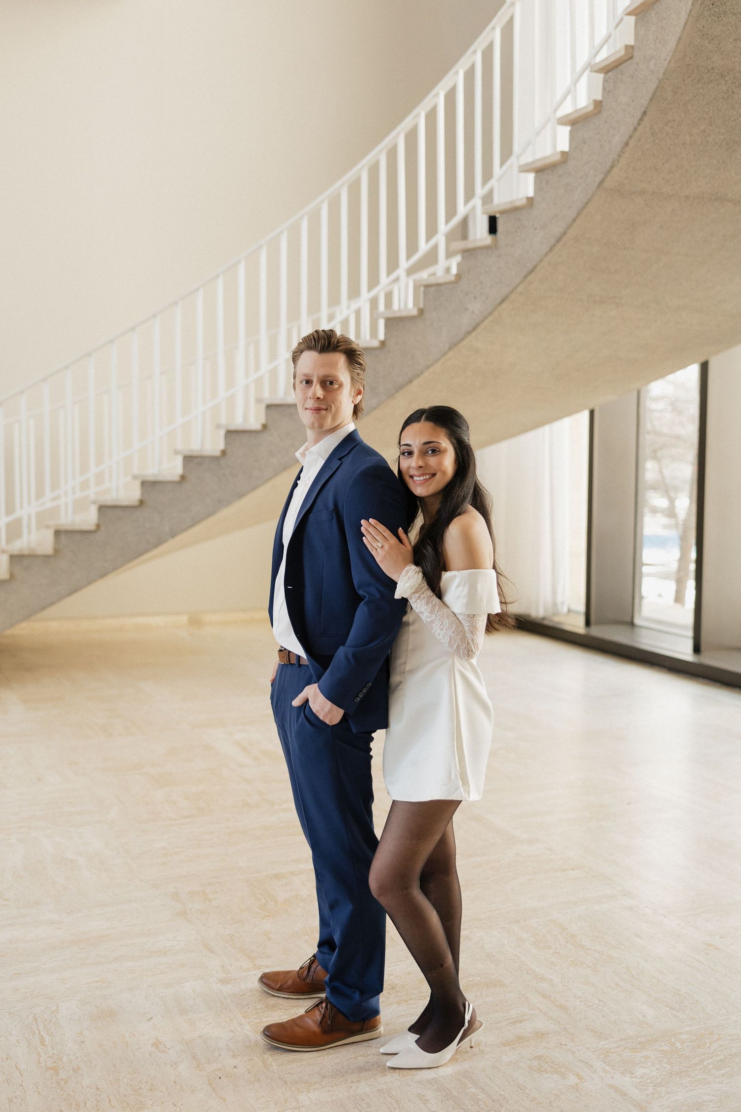 Couple in formal attire posing together near curved white staircase.