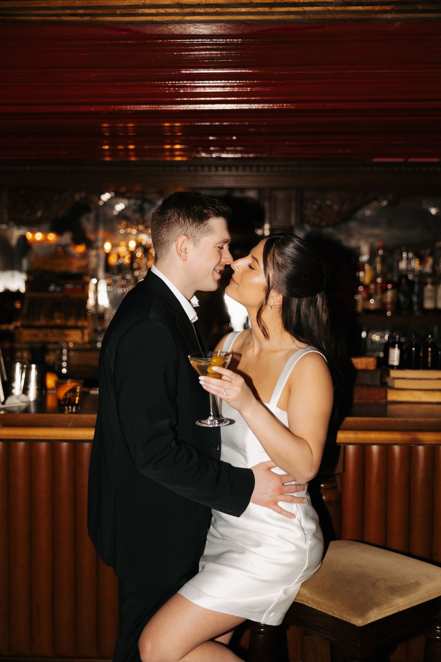 Couple in formal attire embraces while sharing drinks at an upscale bar counter.