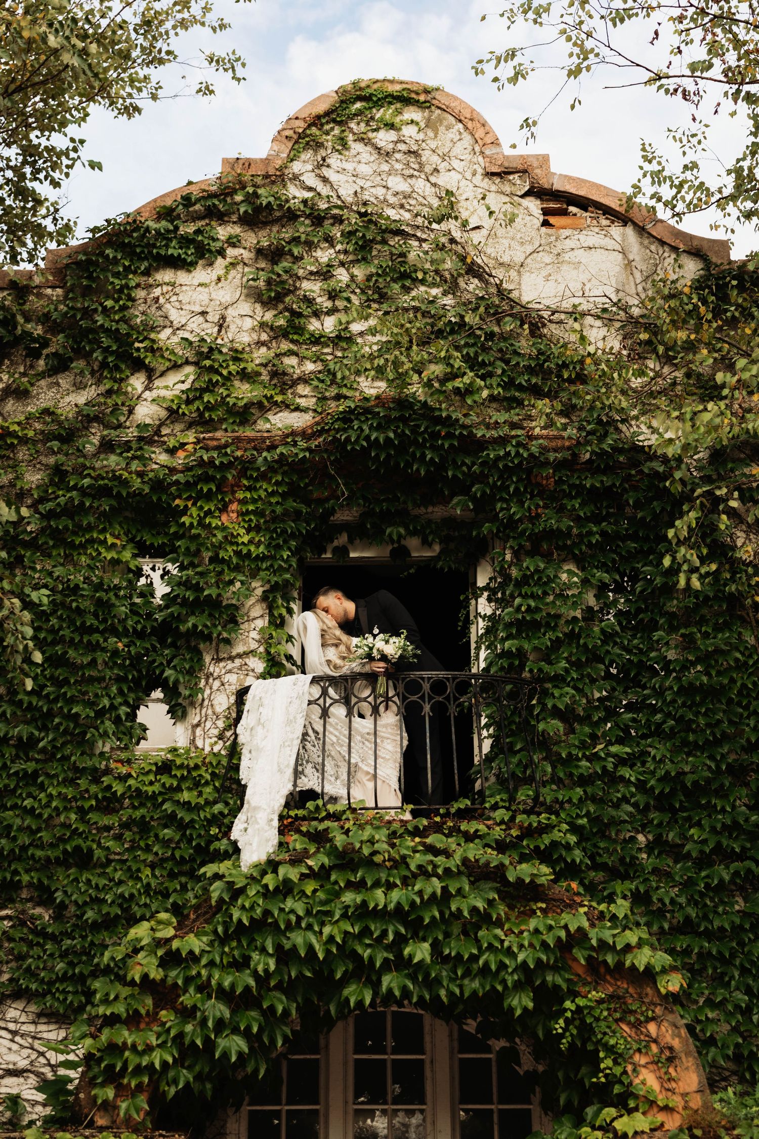 Bride and groom share a kiss on a balcony of an ivy-covered stucco building, her long white veil draping over the railing.