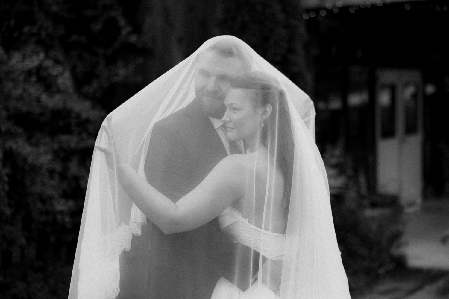 Black and white photo of a bride and groom embracing closely under a sheer wedding veil outdoors.