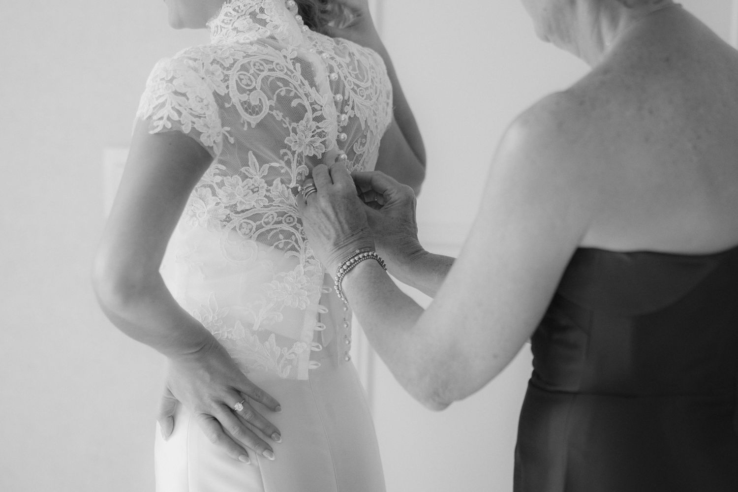 Black and white photo of a woman in a dark dress helping a bride button the back of her lace wedding gown.