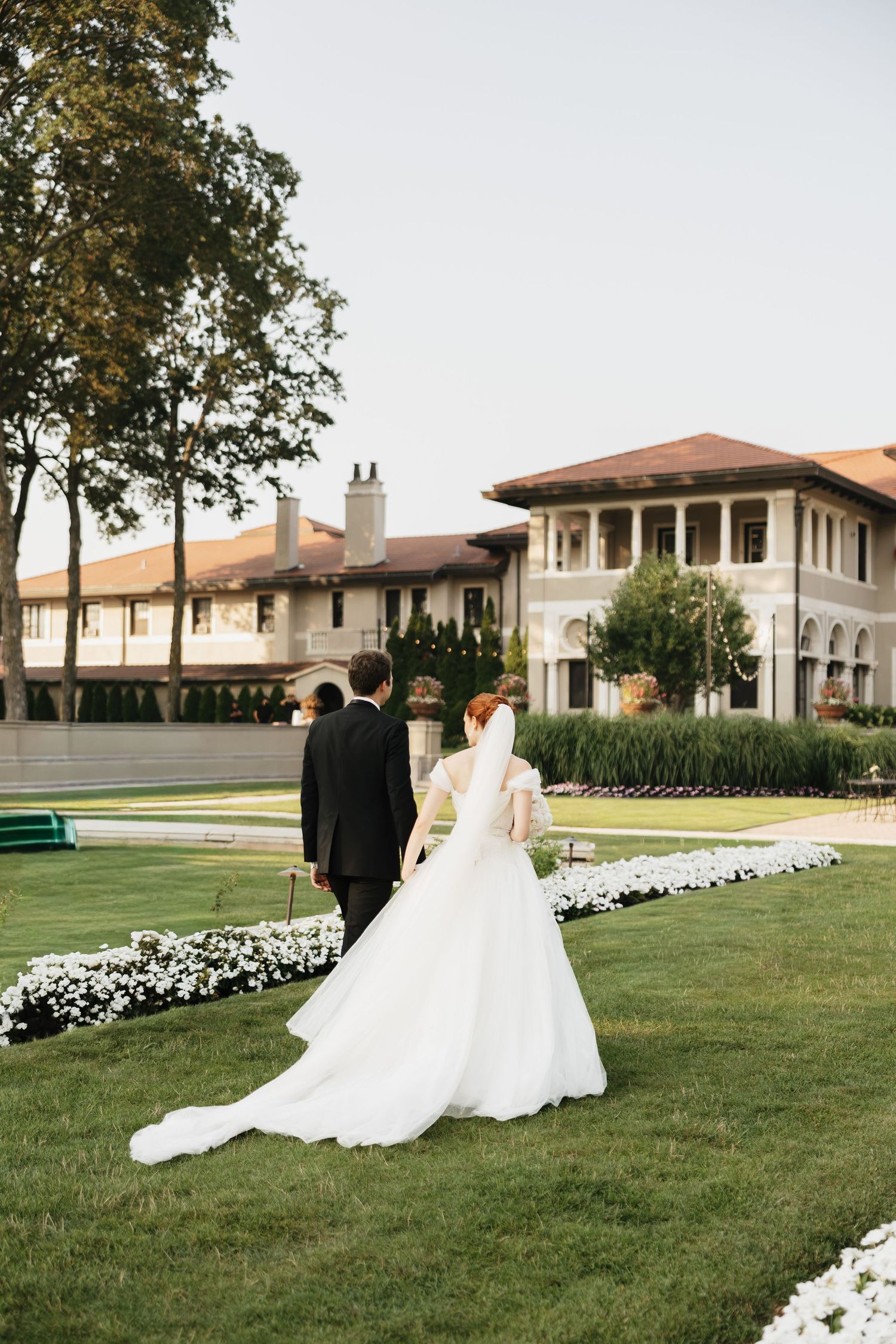 Bride in a flowing white gown and groom in a dark suit walk across a manicured lawn toward an elegant estate at a garden wedding.