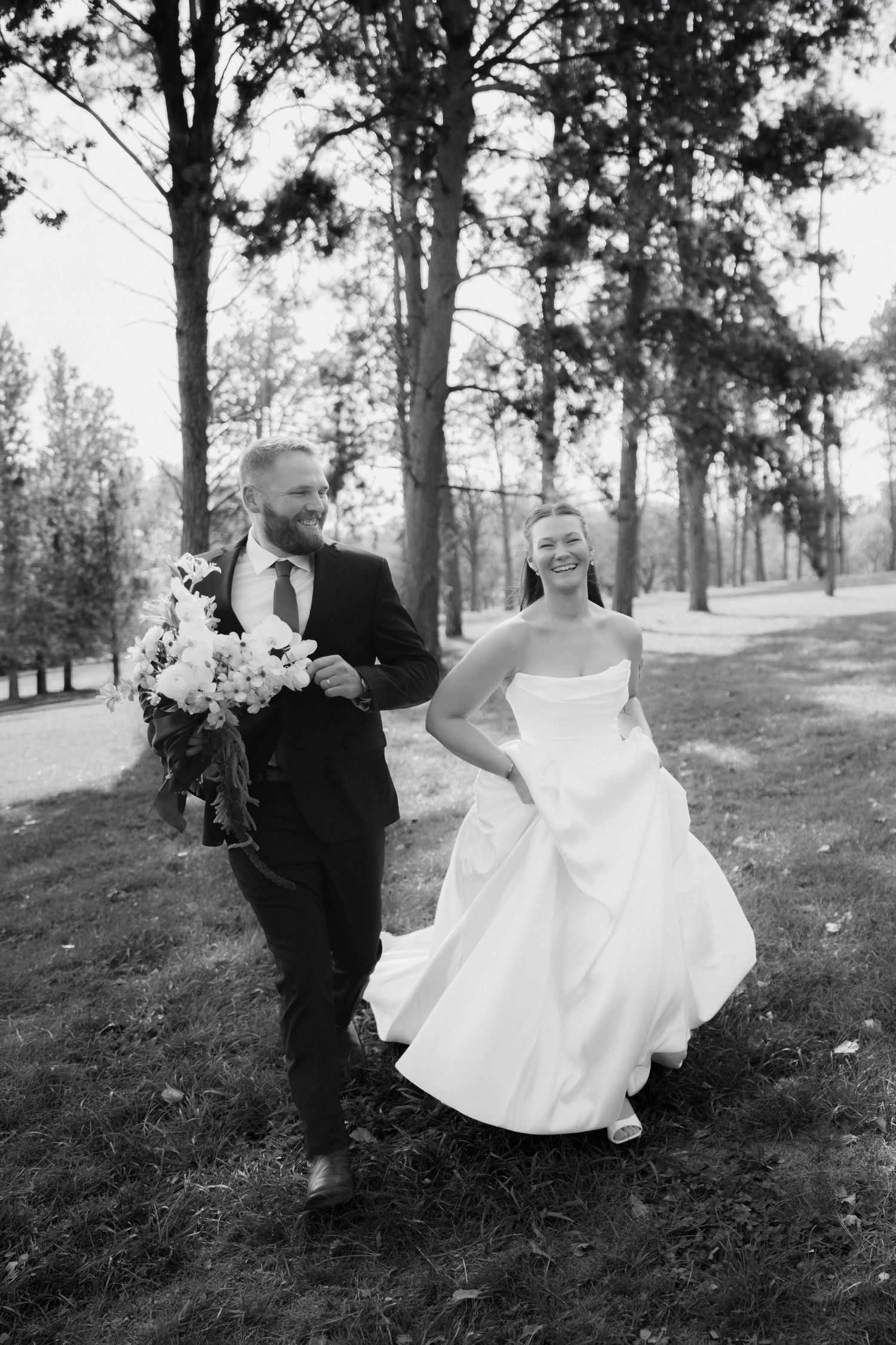 Black and white photo of a laughing bride in a strapless gown walking with a groom in a suit holding a bouquet among tall trees.