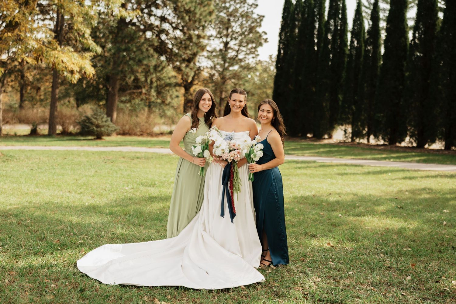 Bride in a white strapless gown holding a bouquet poses with two bridesmaids in green dresses on a sunny lawn framed by trees.