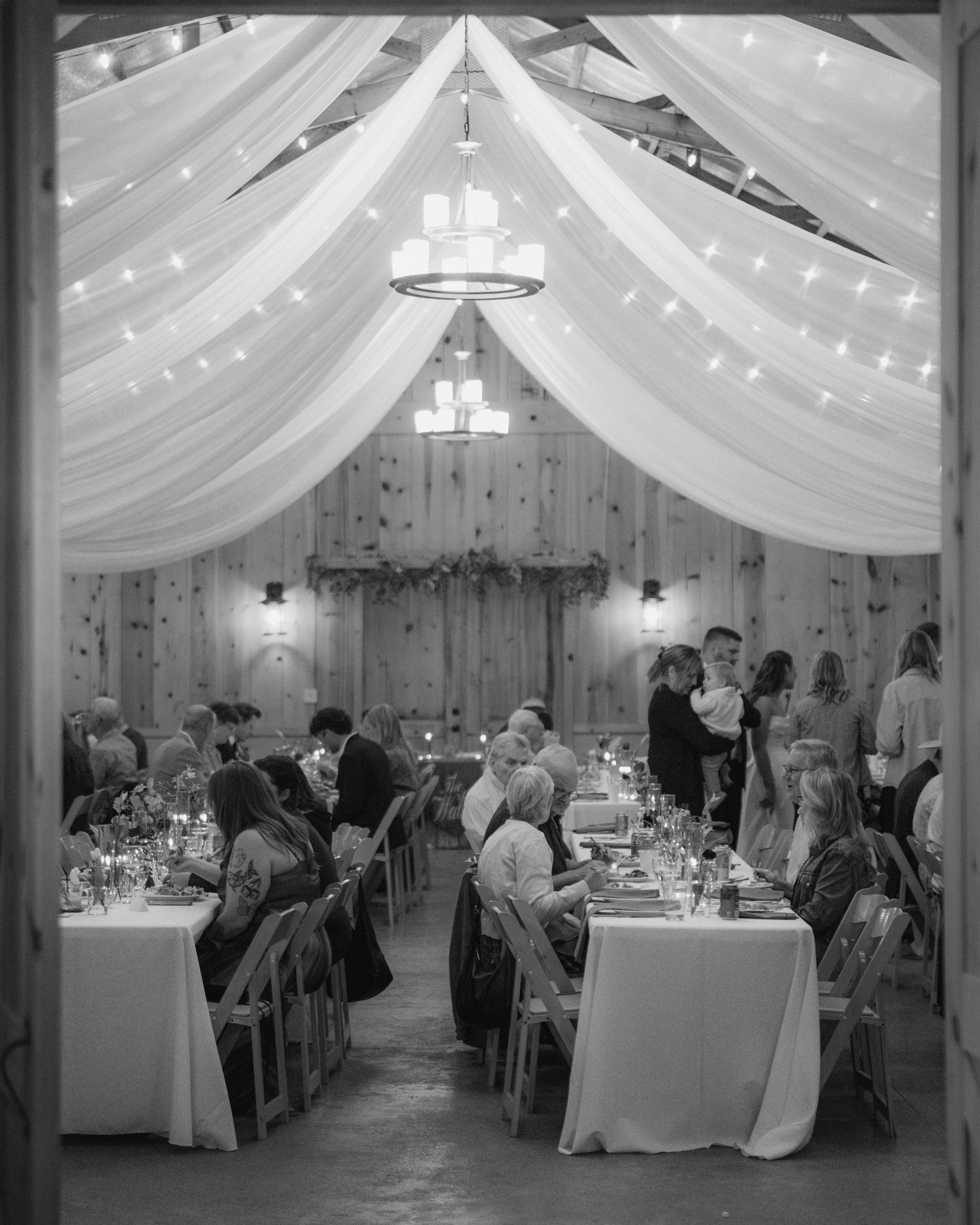 Black and white photo of wedding guests dining at long tables in a rustic barn with draped fabric ceiling, string lights, and chandeliers.