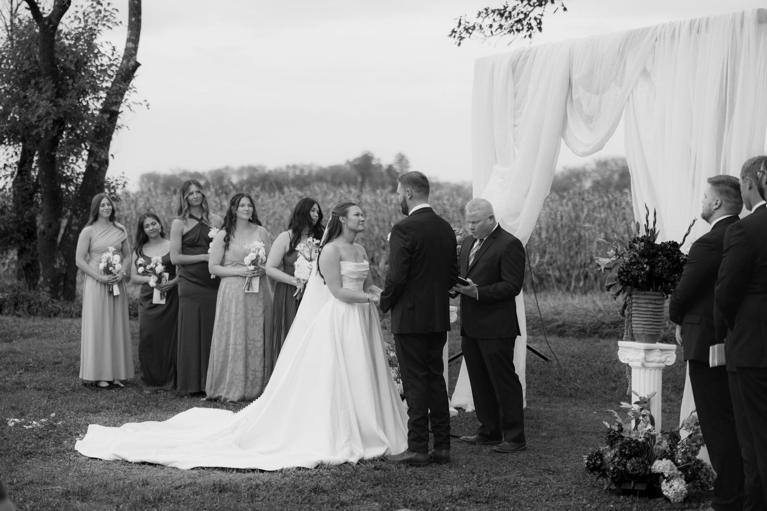 Black and white outdoor wedding ceremony with a bride in a long gown, groom, officiant, and bridesmaids standing beside a draped altar.