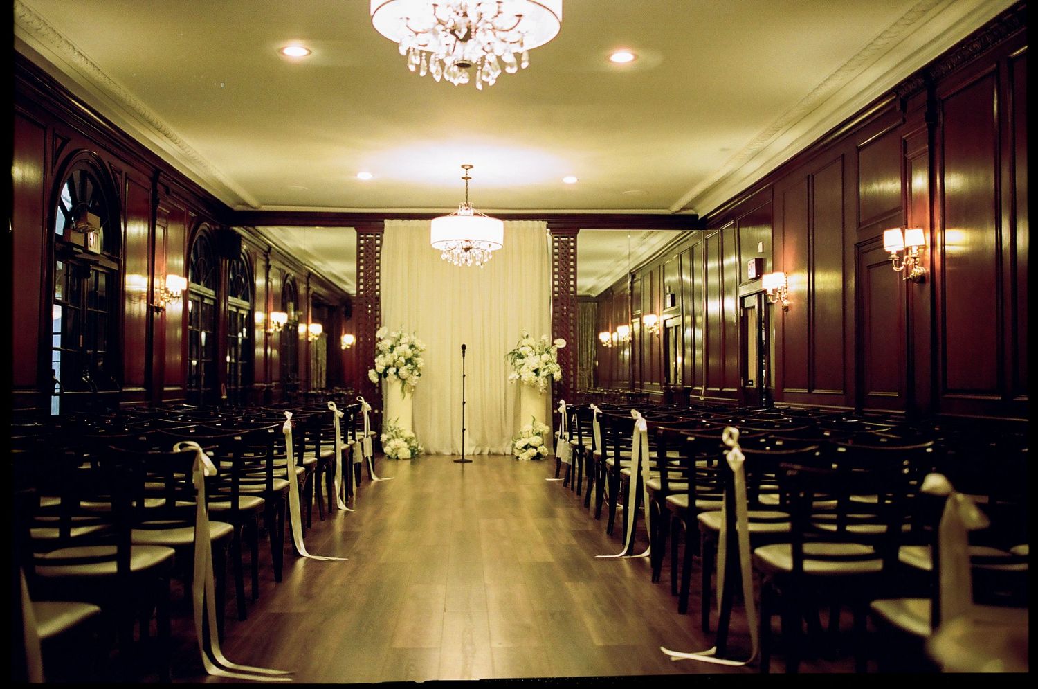 Elegant wedding ceremony setup in a dark wood-paneled ballroom with chandelier lighting and white floral altar.