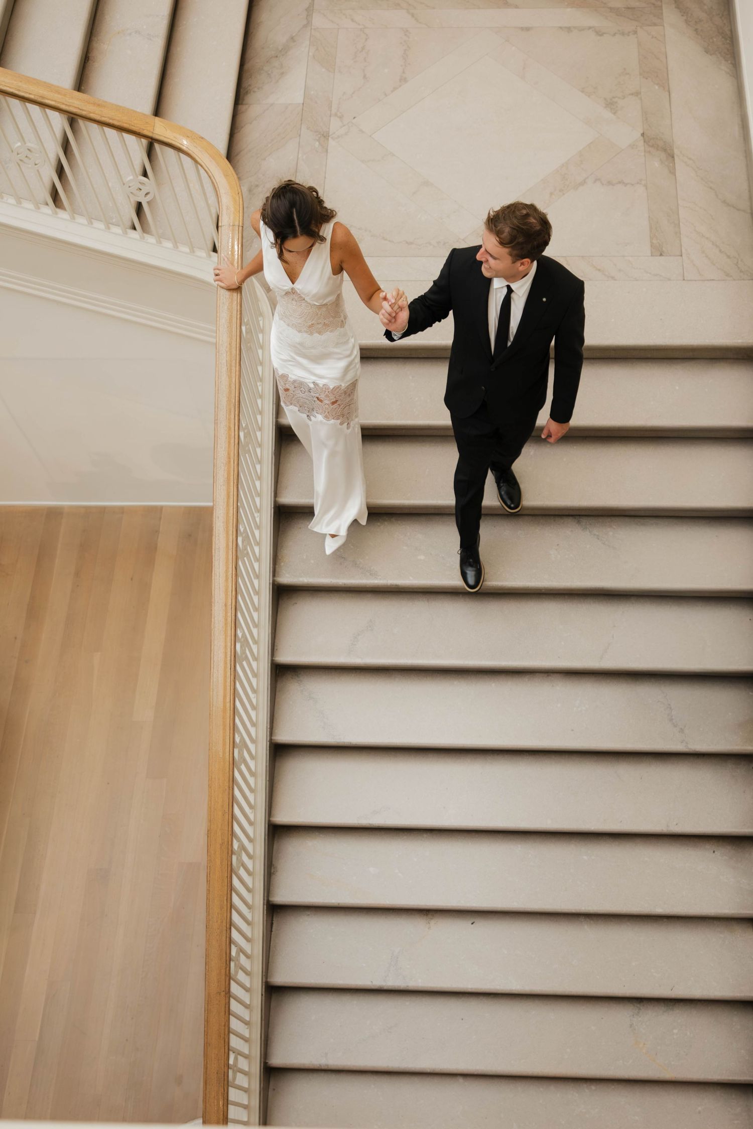 Aerial view of bride and groom holding hands walking up marble stairs, bride in white gown, groom in black suit.