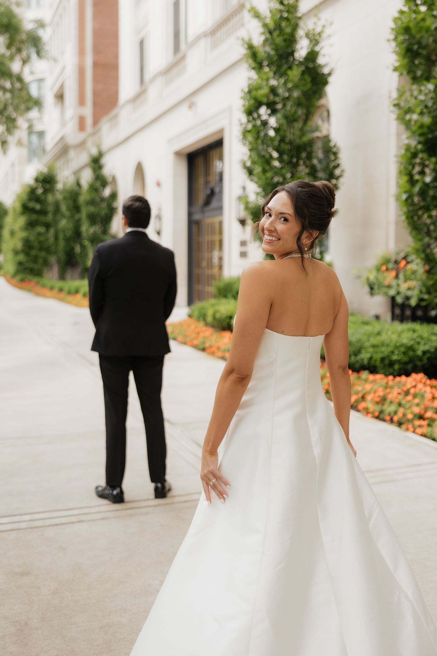 Bride in strapless white gown smiles over shoulder while groom in black tuxedo stands ahead on elegant outdoor walkway.