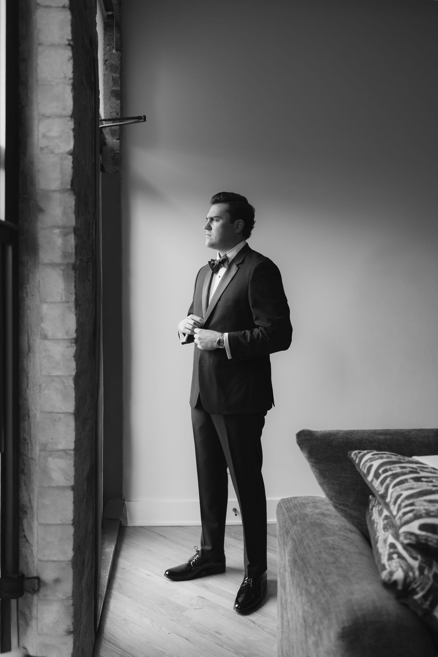 Black and white photo of an elegant groom in a tuxedo adjusting his cufflinks by a window in a stylish room.