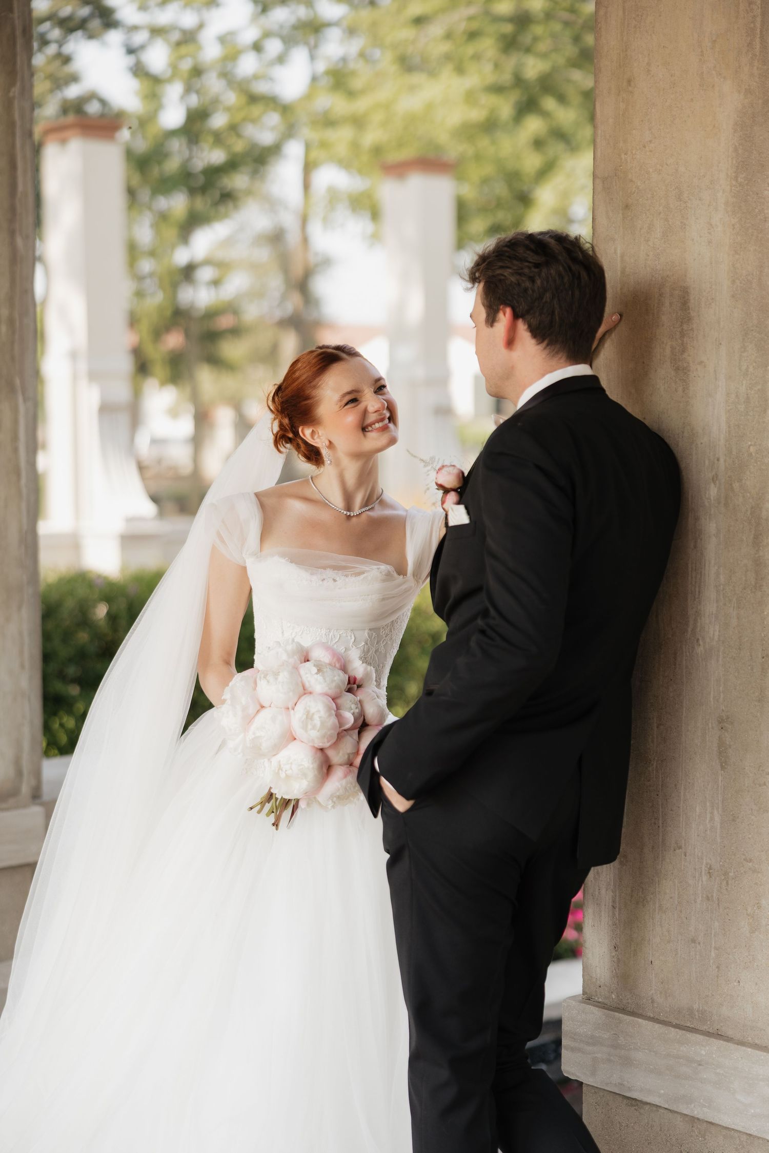 Smiling red-haired bride in white gown holds pink peony bouquet while standing with groom by stone columns.