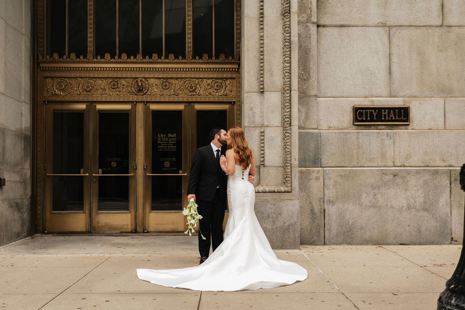 Bride and groom share a romantic kiss outside City Hall, bride in elegant white gown with long train.