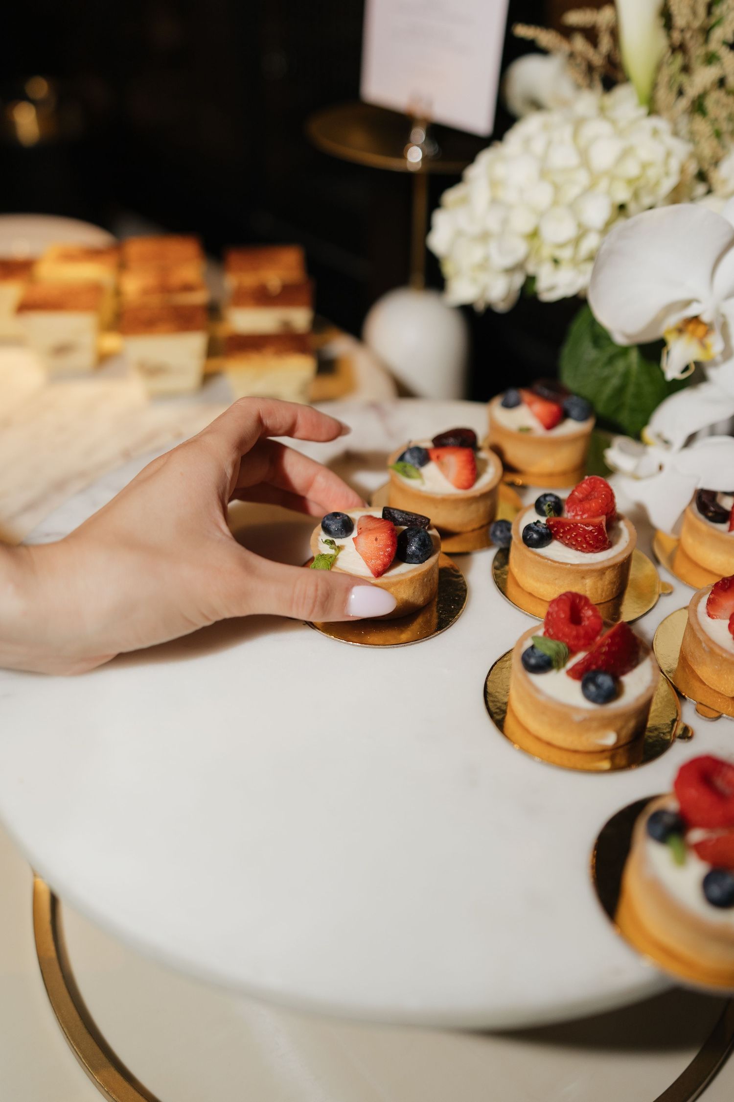 A hand picks a mini fruit tart topped with strawberries and blueberries from a dessert display at a wedding reception.