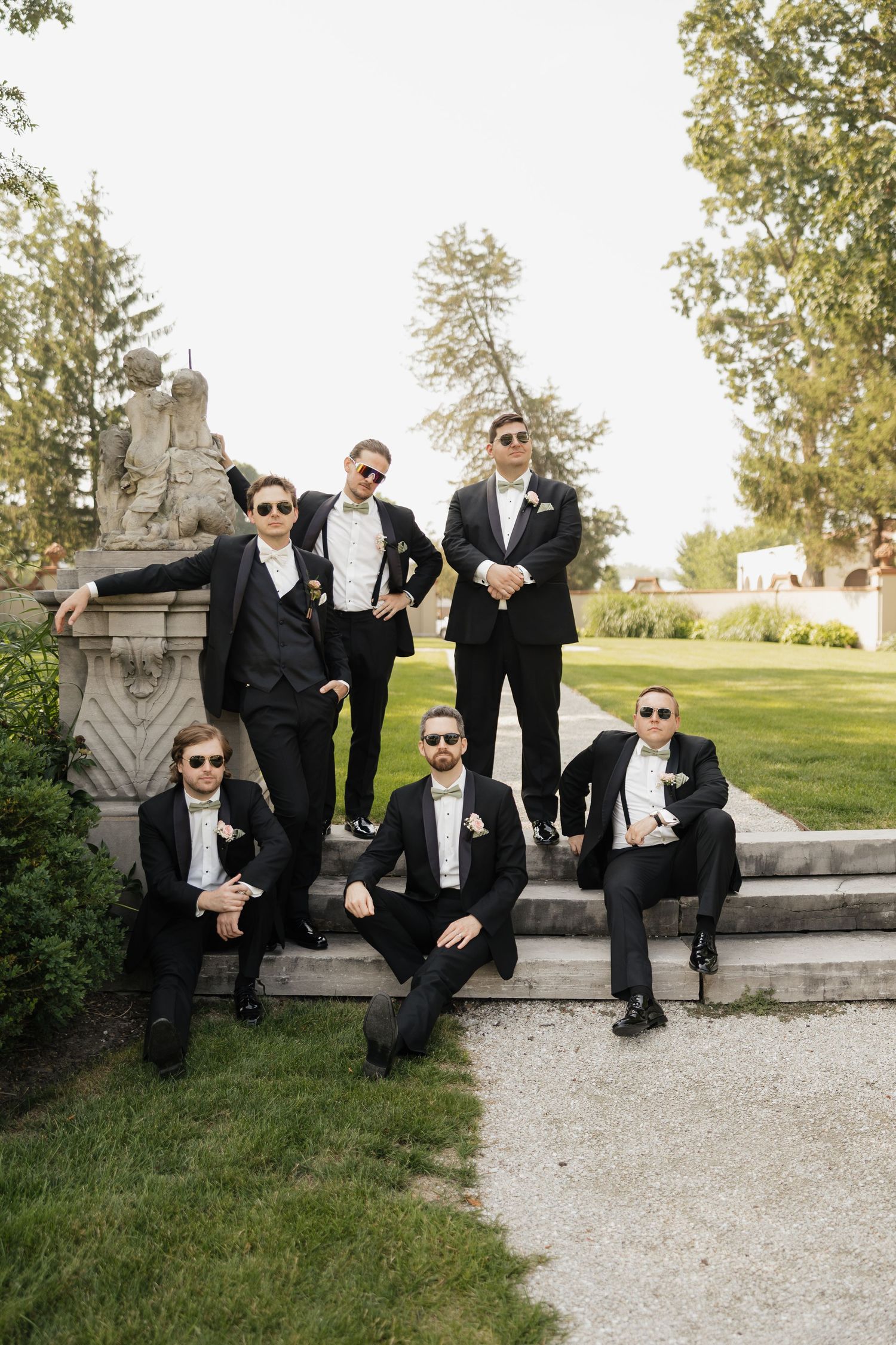 Five groomsmen in black tuxedos and sunglasses posing on stone steps beside classical garden sculpture.