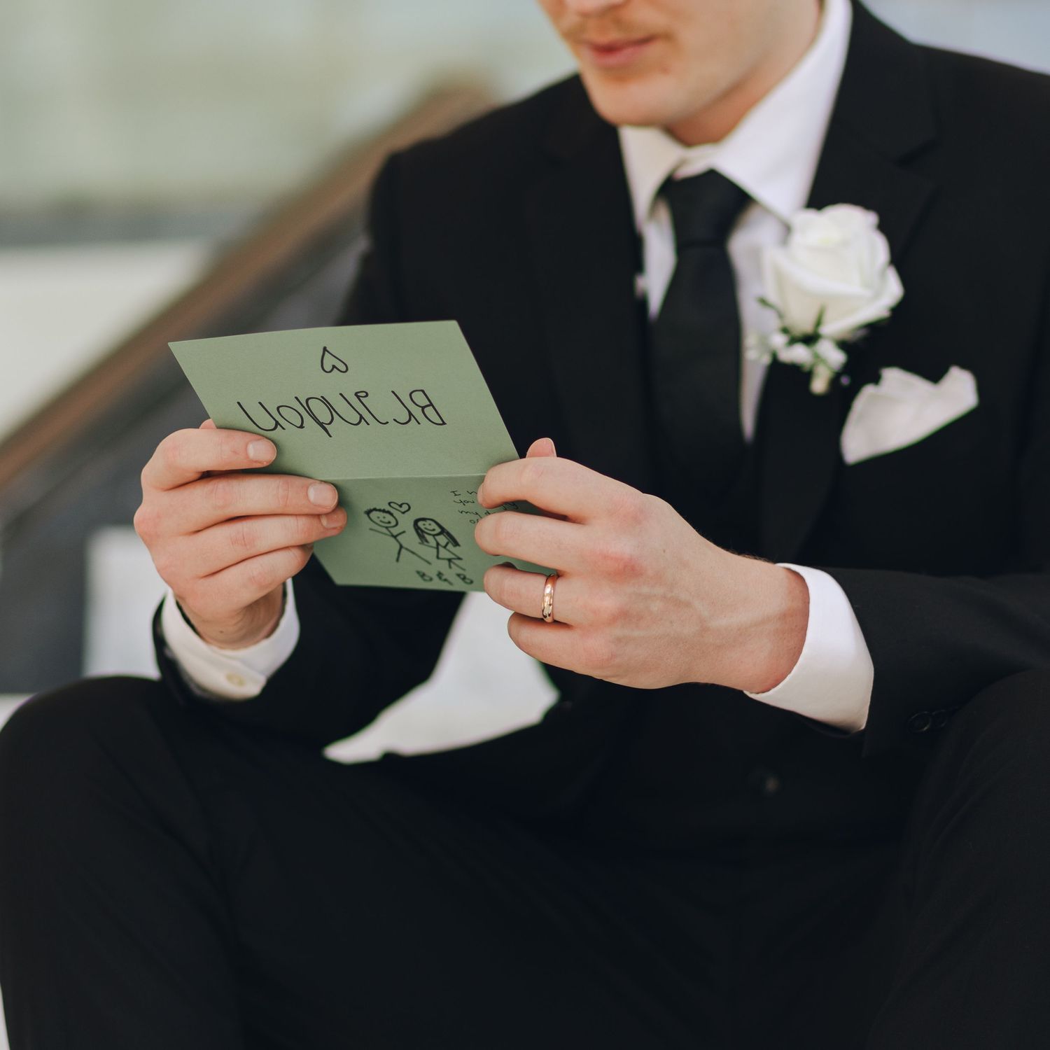 A groom in black suit with white boutonniere reads a handwritten note on green paper during wedding preparations.