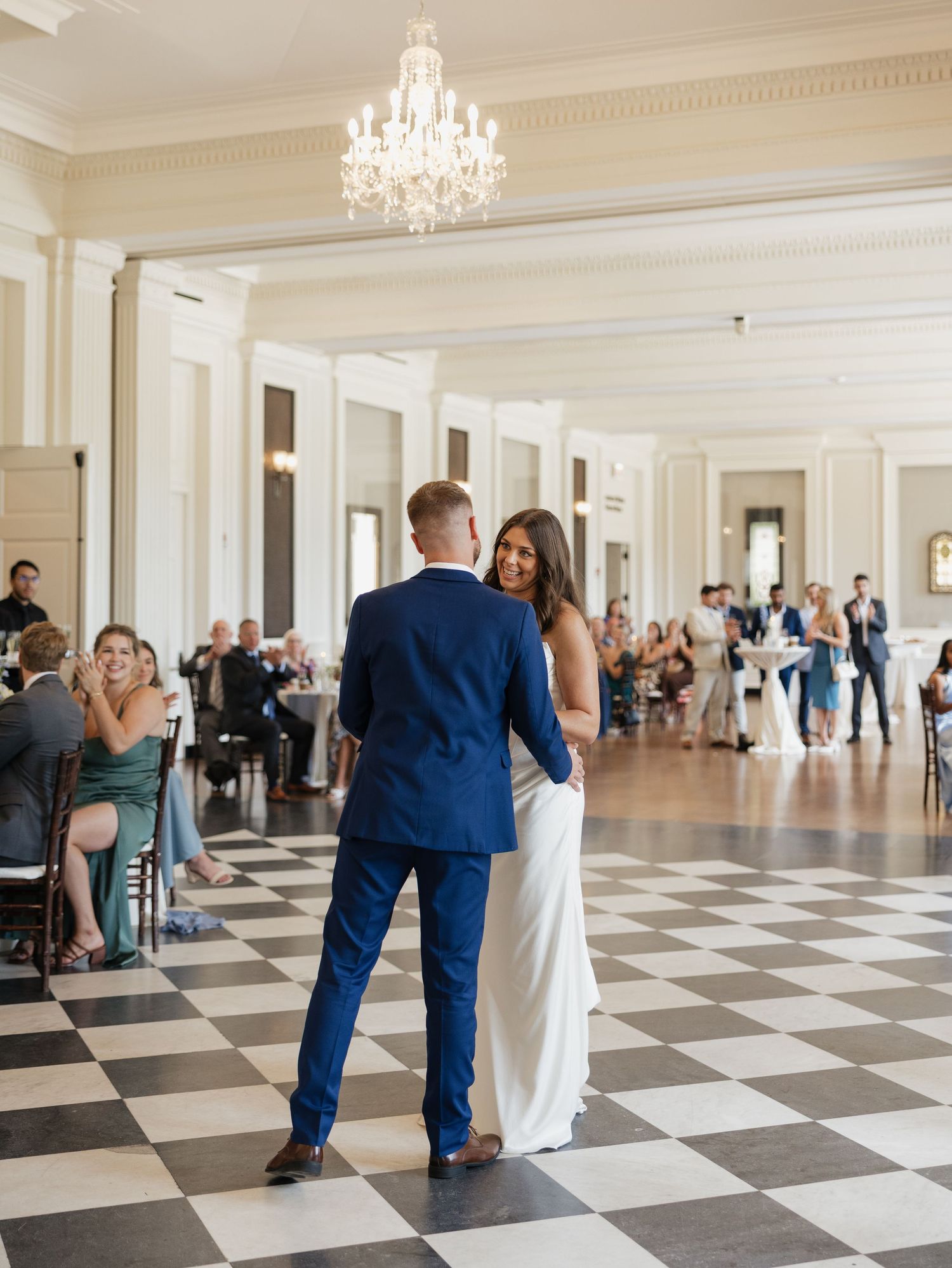 A couple dances together in an elegant ballroom with black and white checkered floors and crystal chandeliers.