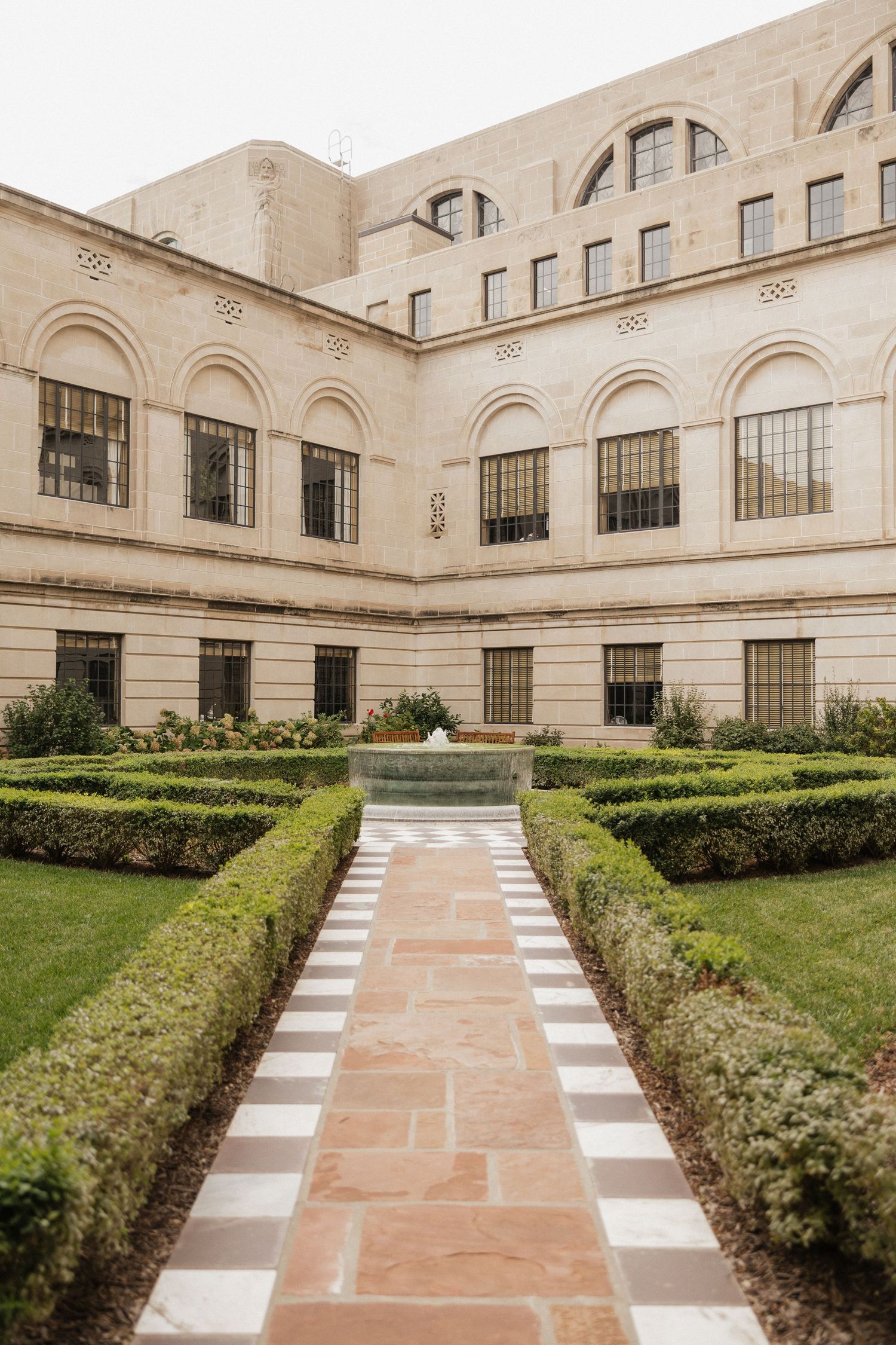 A formal garden path with stone pavers and manicured hedges leads to a grand beige building with arched windows.