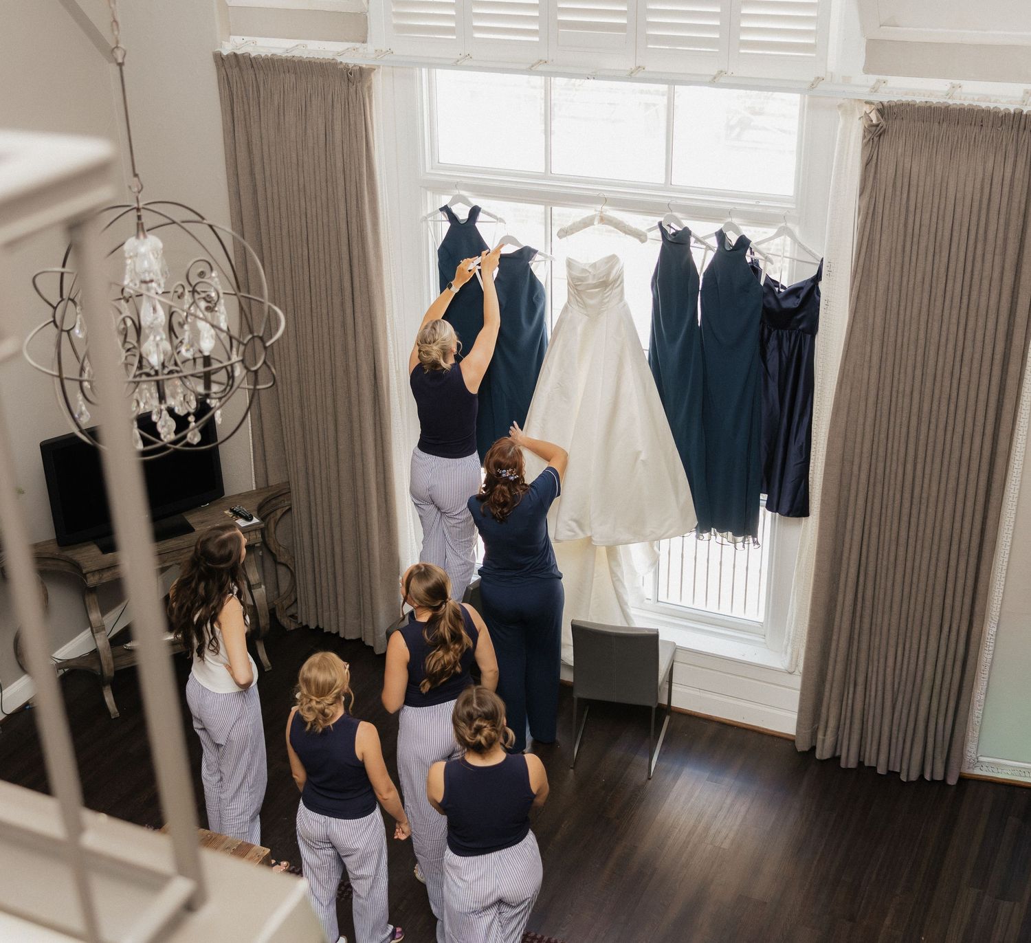 Bridesmaids in navy dresses gather around a window where a white wedding gown and dark bridesmaid dresses hang elegantly.