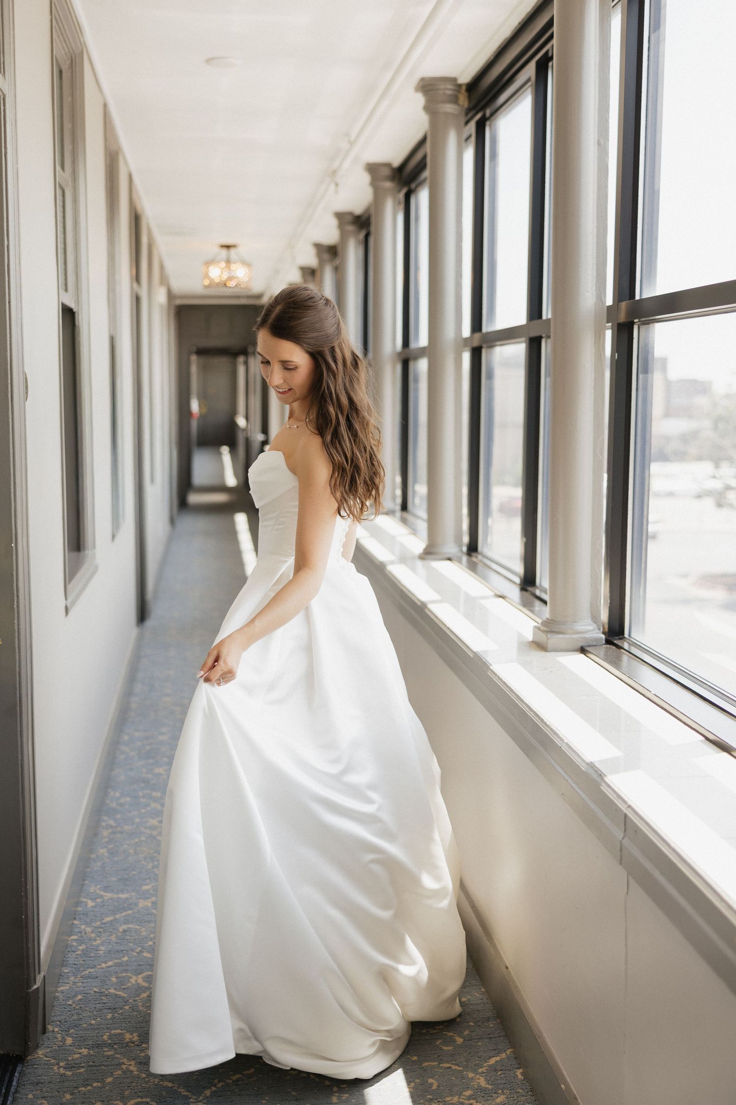 Bride in a strapless white ballgown standing in a sunlit hallway with large windows, holding her dress elegantly.