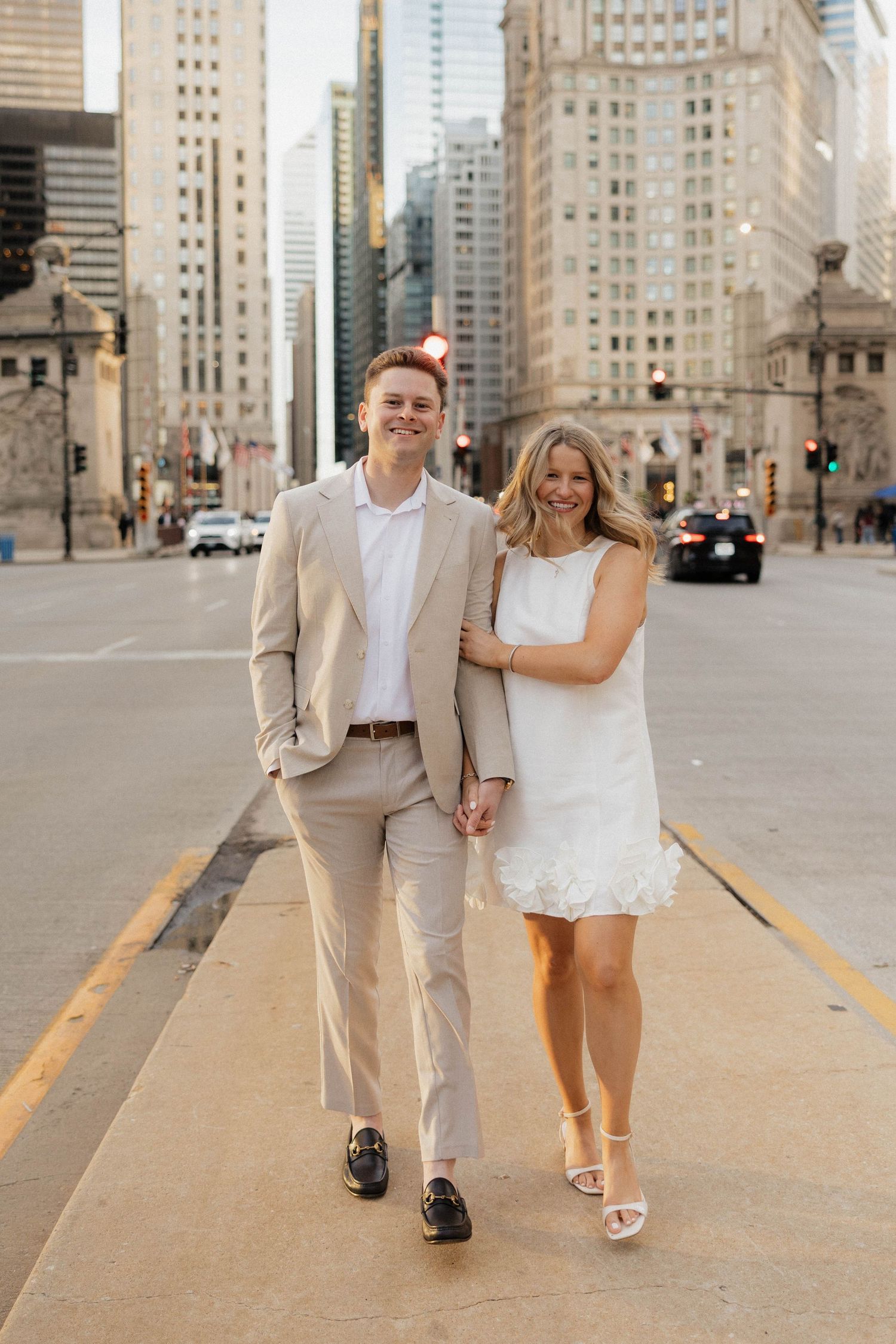 Couple walks hand-in-hand in Chicago, man in tan suit, woman in white dress, city skyscrapers in background.