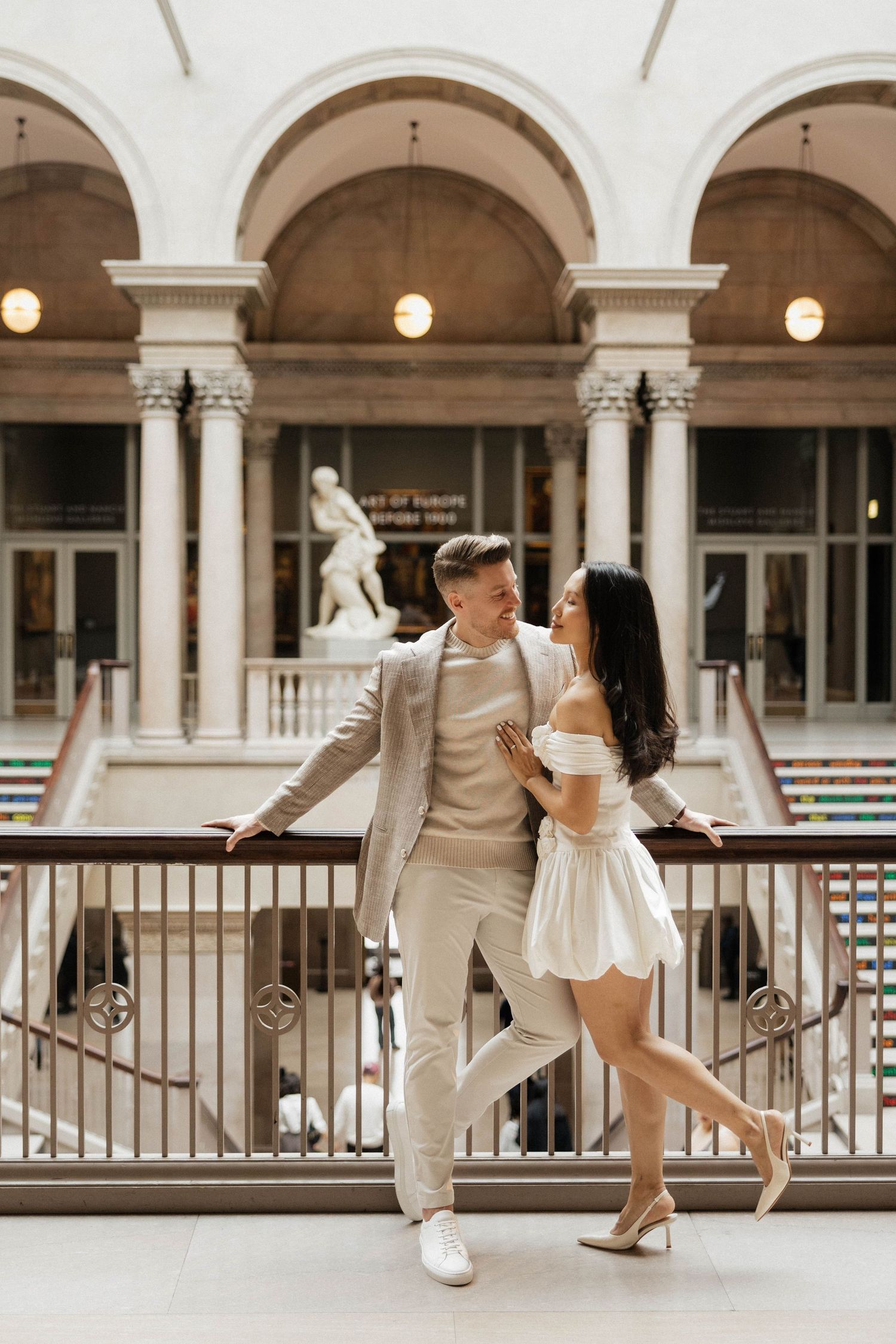 Elegant couple in white outfits posing at a grand museum balcony with classical arches and sculpture.