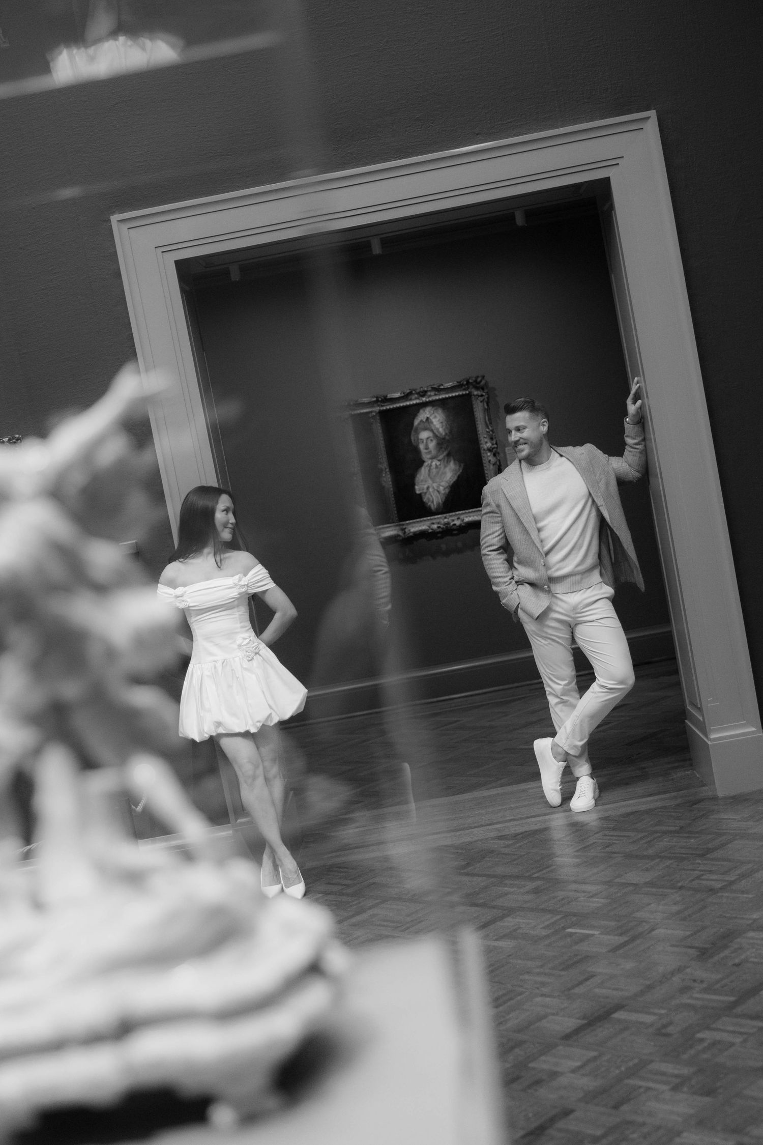 Black and white photo of a stylish couple walking through an art museum gallery with a classic portrait painting in the background.