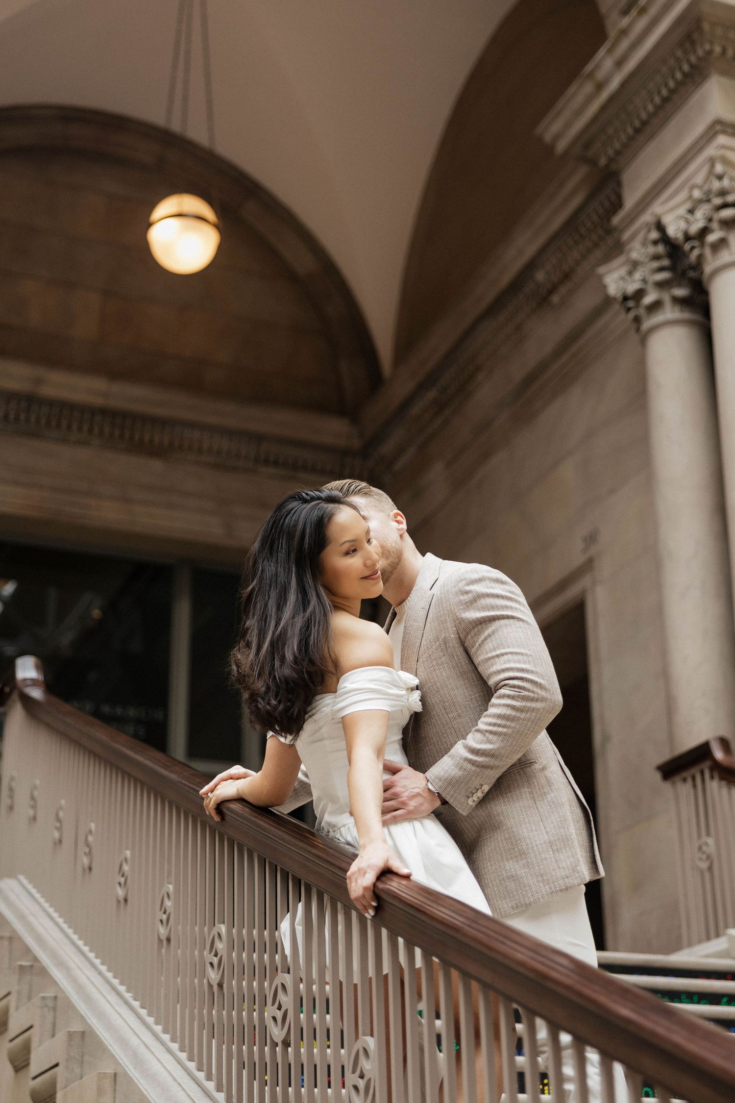 Couple in wedding attire sharing a romantic moment on elegant staircase with arched stone architecture.
