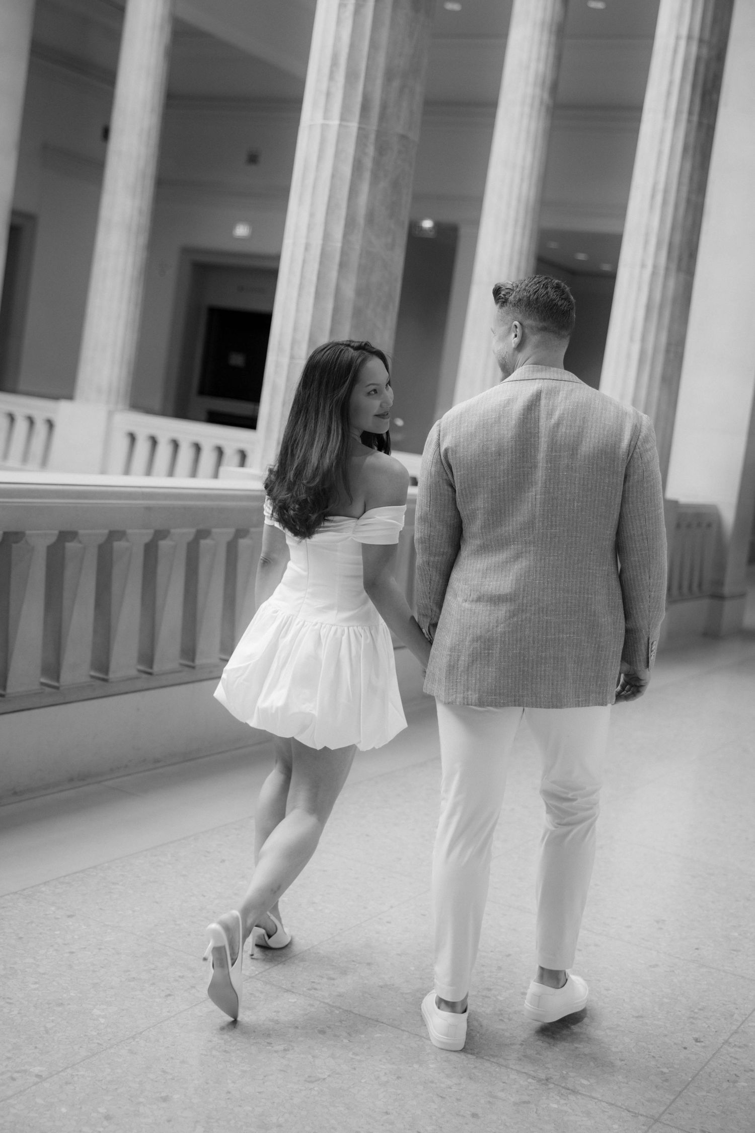 Black and white photo of a couple walking hand in hand through elegant columned hall, woman in white mini dress glancing back.