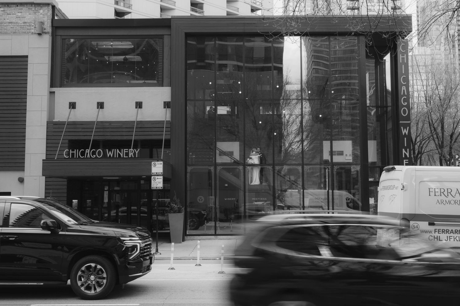 Black and white street photo of Chicago Winery exterior with glass facade and cars passing by.