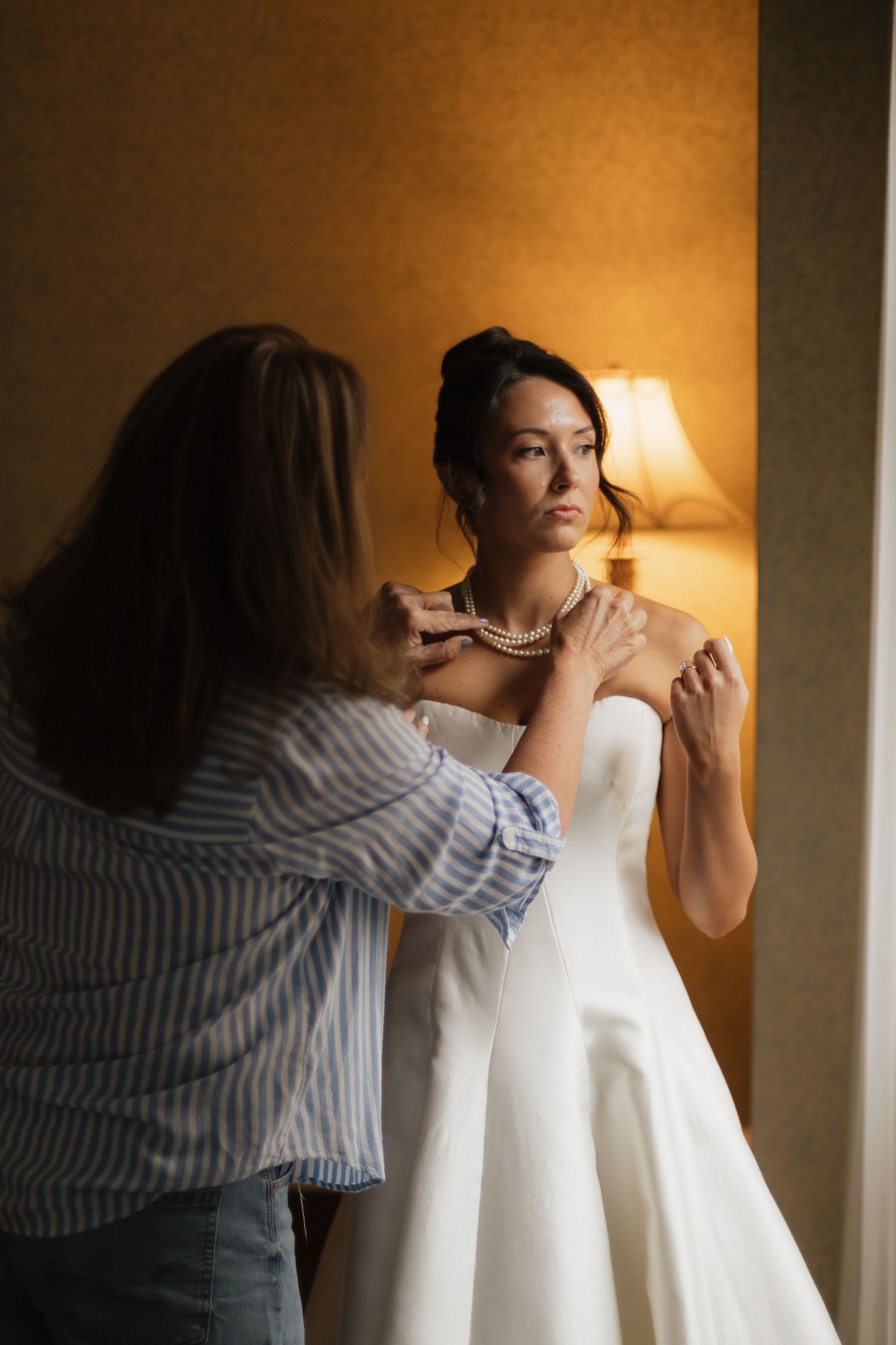 A bridesmaid fastens a pearl necklace on a bride in a white strapless gown, lit by a warm lamp.
