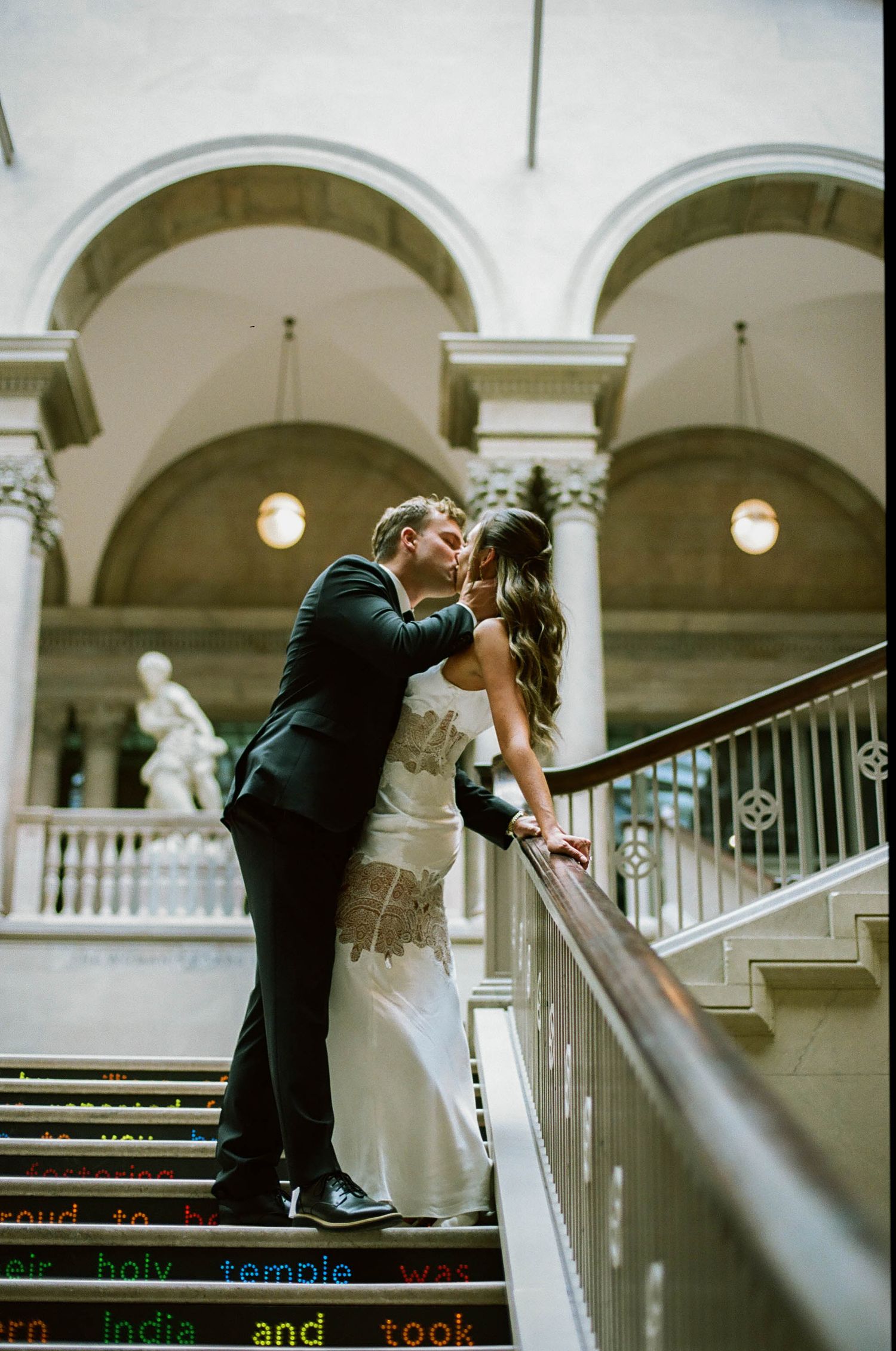 Bride and groom sharing a romantic kiss on an elegant staircase with arched ceilings in a grand venue.