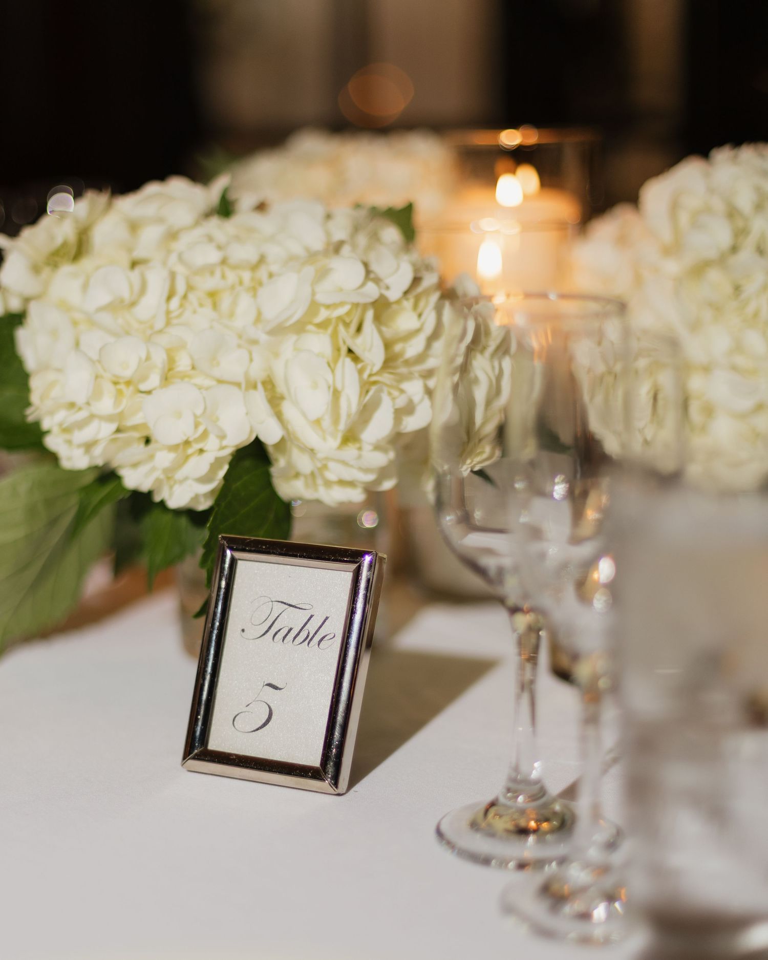 Elegant wedding table centerpiece with white hydrangeas, silver-framed Table 5 number card, crystal glasses, and glowing candles.