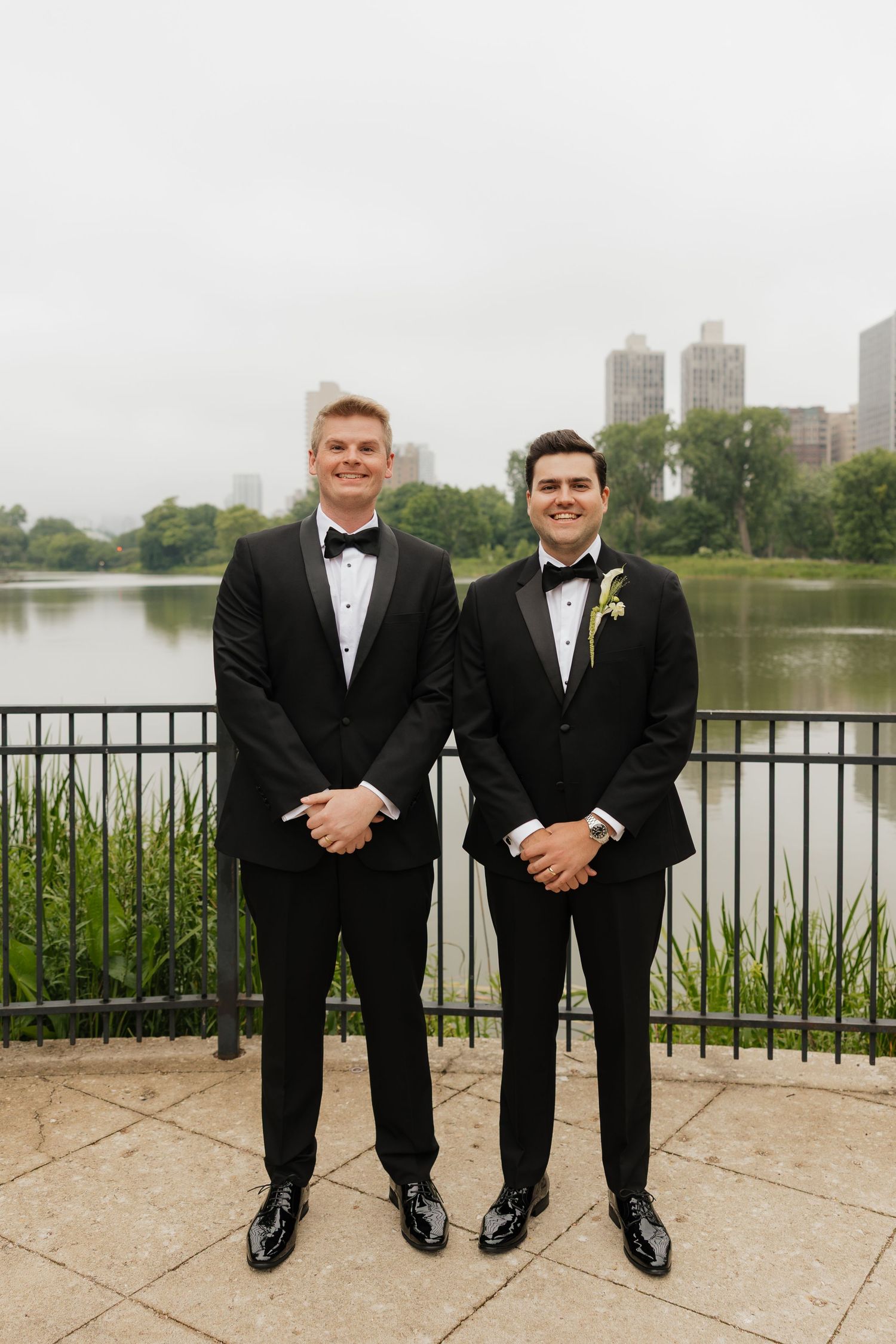 Two grooms in black tuxedos with bow ties pose smiling by a lakeside railing with city skyline backdrop.