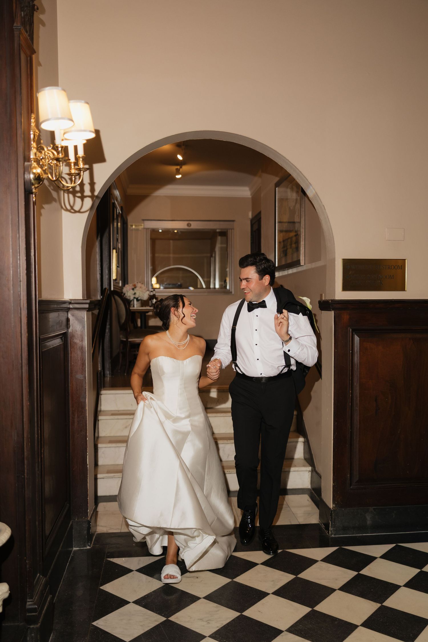 Bride and groom walk hand-in-hand through elegant arched doorway on black and white checkered floor.