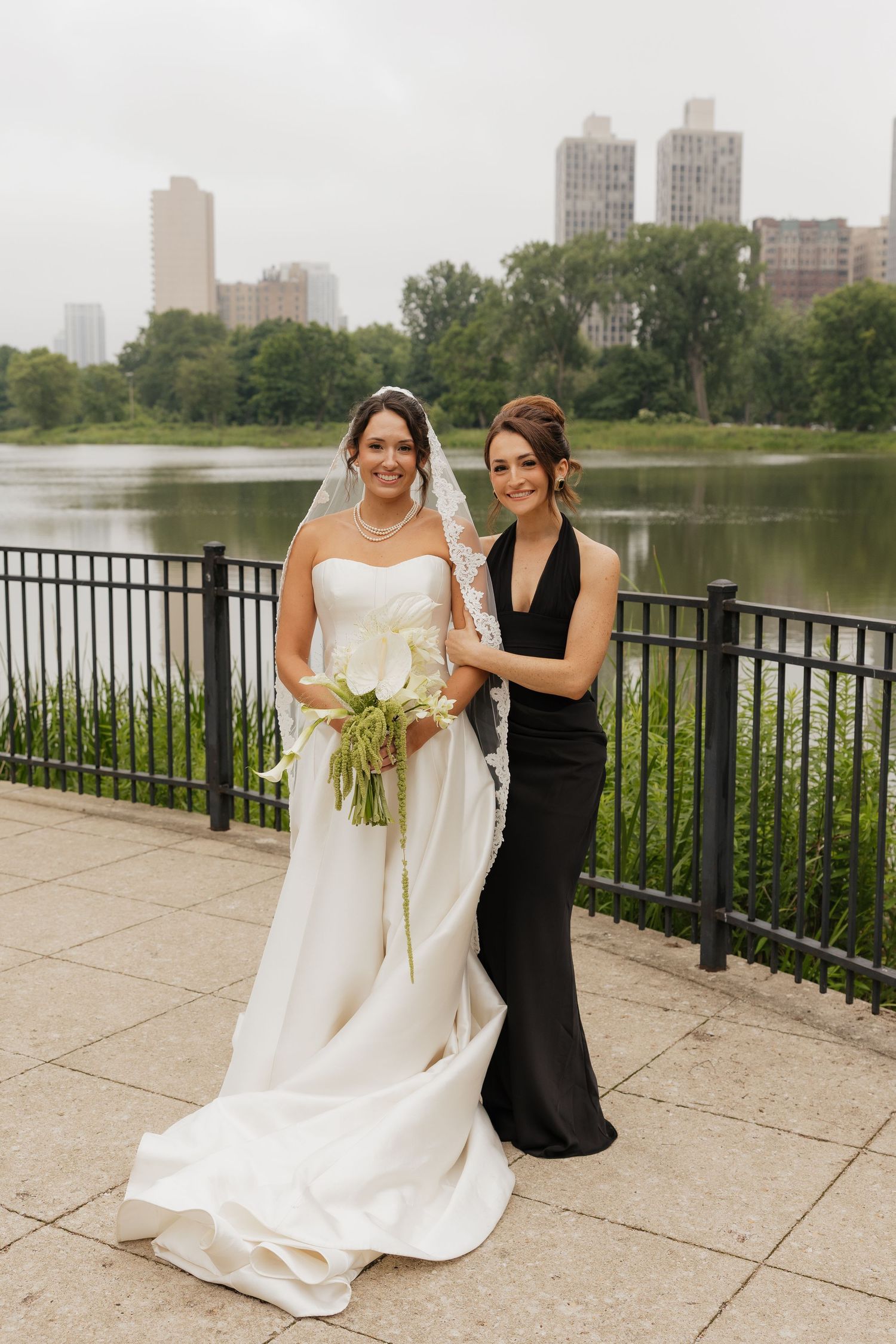 Bride in white gown with veil holds bouquet beside bridesmaid in black dress, posing by a lake in Central Park, NYC.