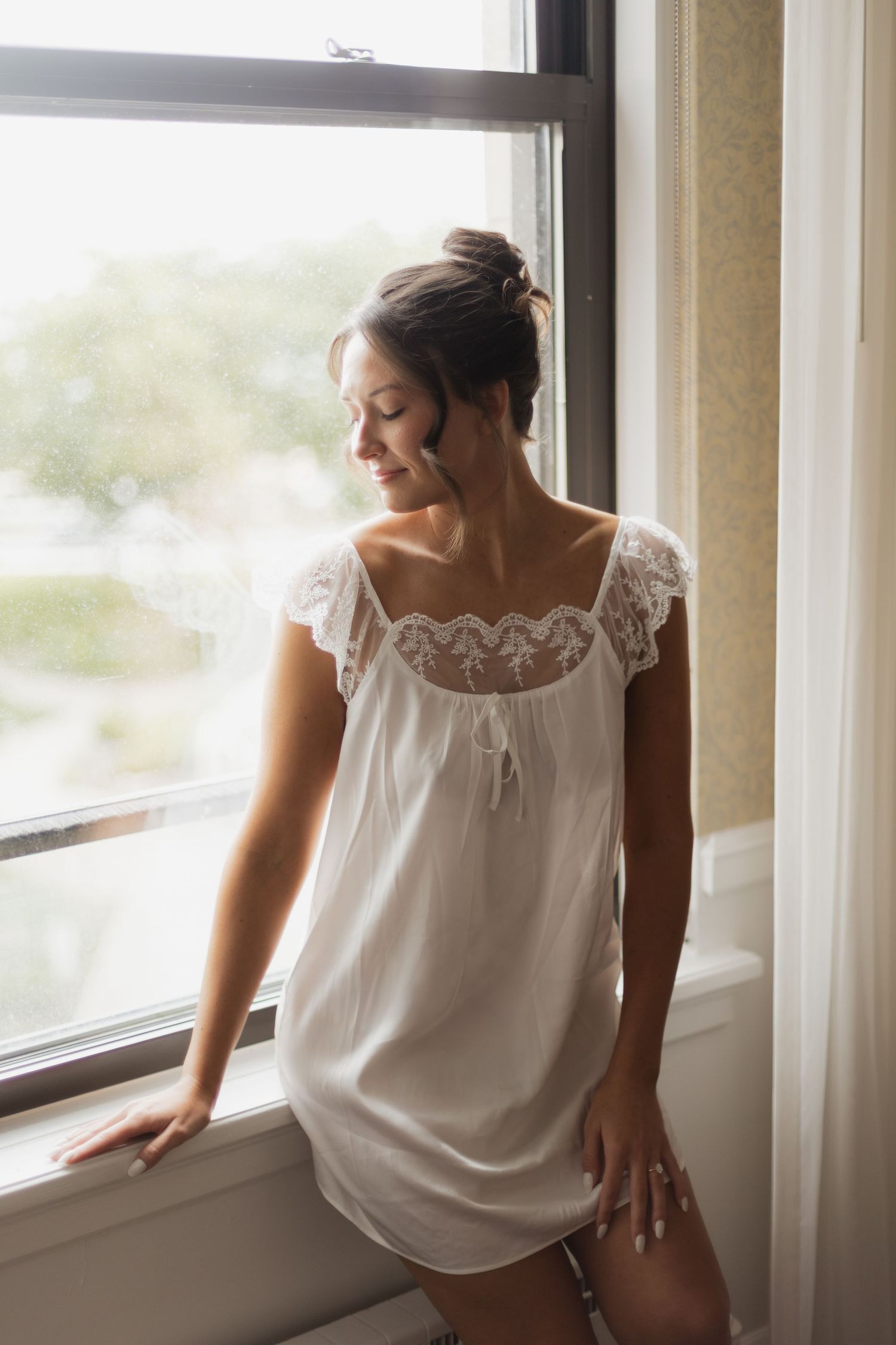 Woman in white lace chemise nightgown standing by a sunlit window, looking down gracefully.