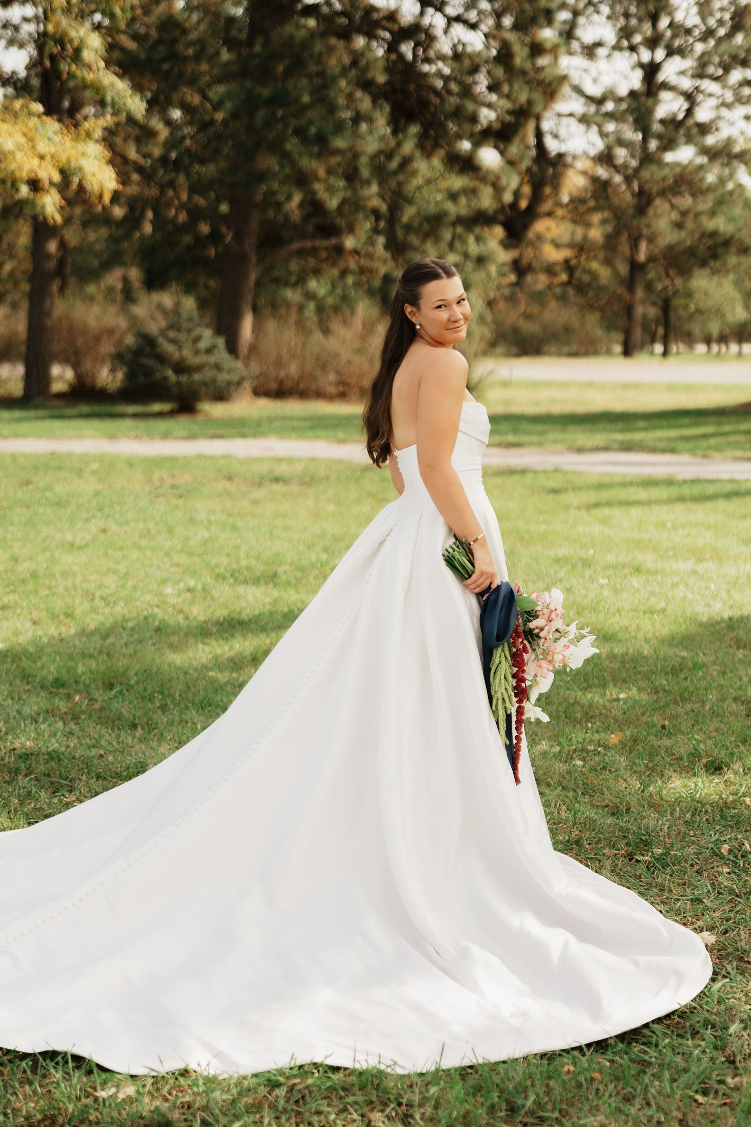 Bride in a strapless white satin wedding gown with a long train holds a bouquet while smiling on a sunny green lawn surrounded by trees.