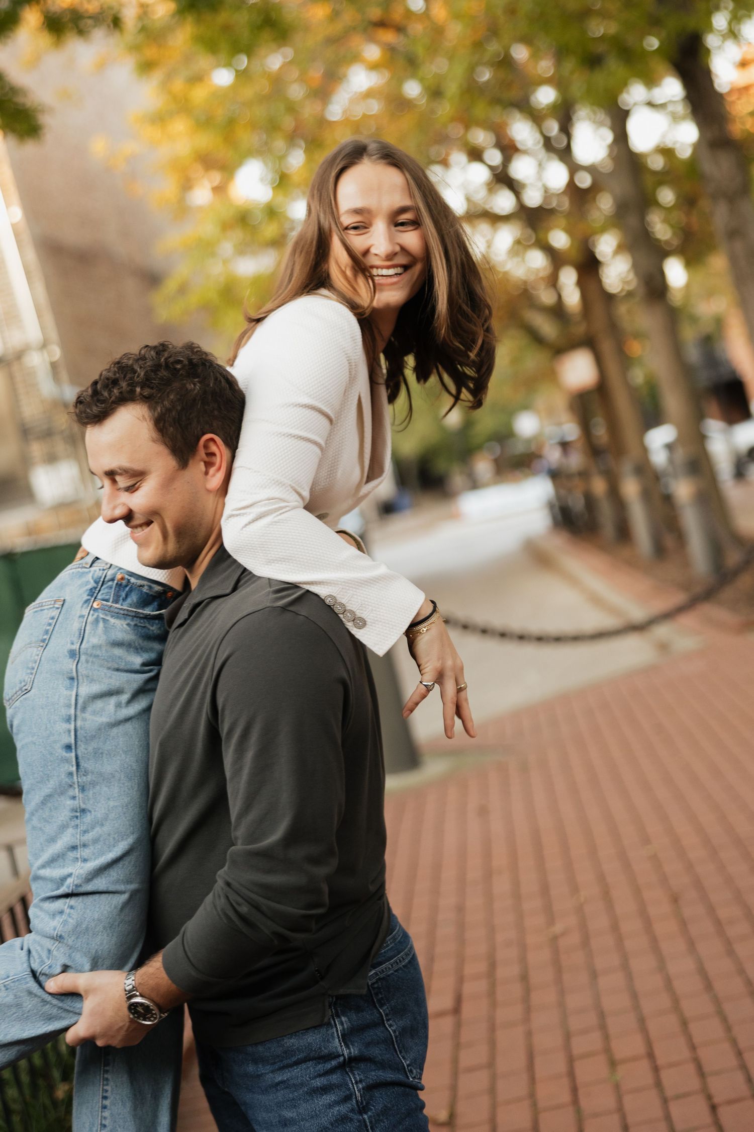 A smiling couple embraces outdoors on a brick sidewalk as a woman in a white top leans playfully over a man's back in warm autumn light.