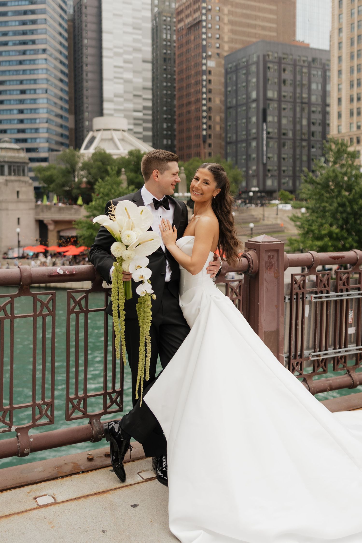 Smiling bride in a strapless white gown and groom in a tuxedo hold a cascading white calla lily bouquet on a city riverwalk bridge.