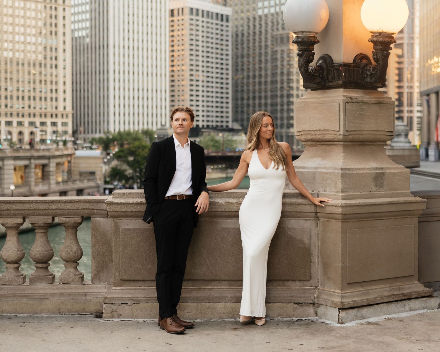 Couple posing at a stone balustrade overlooking a city river, he in a black suit and she in a white dress, with skyscrapers behind them.