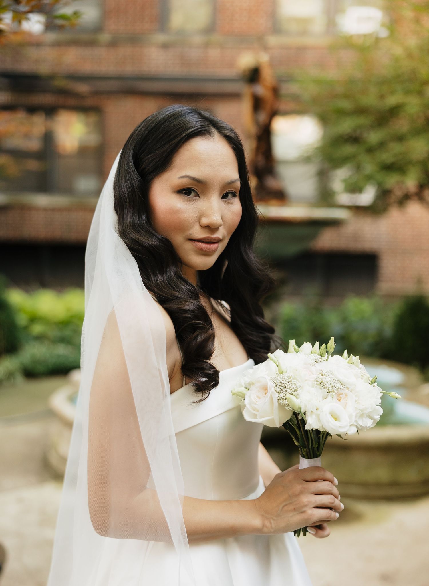 Bride in an elegant white strapless gown and sheer veil holds a bouquet of white roses in a sunlit courtyard garden.