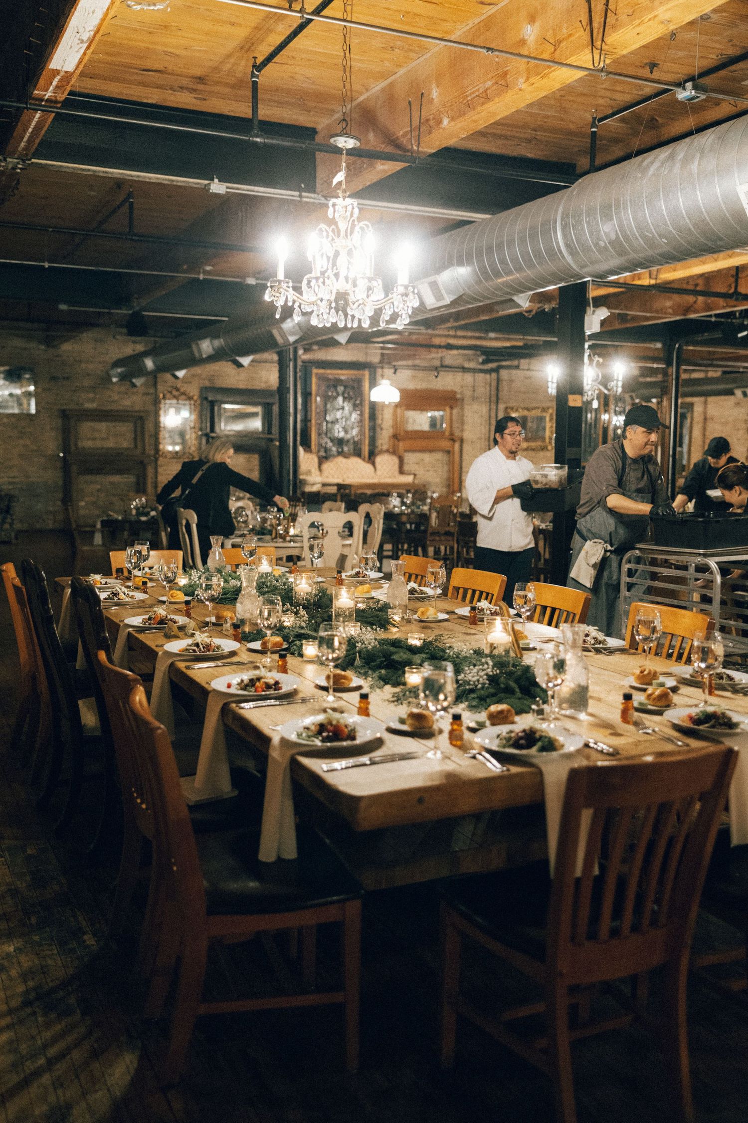 Long wooden dining tables set for an elegant dinner party in an industrial-style space with exposed beams and chandeliers.