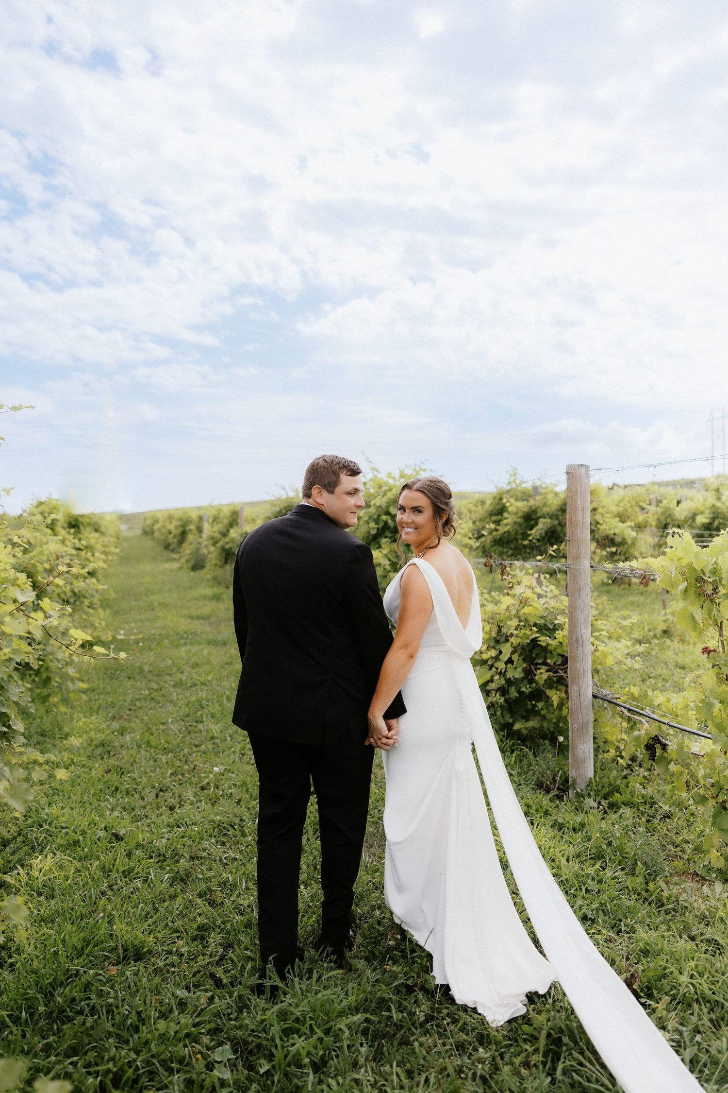 A couple in wedding attire poses together on a grassy hillside with vineyard rows in the background.