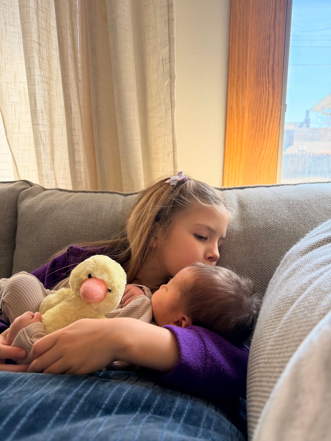 A young girl tenderly kisses a sleeping baby on a gray sofa, holding a yellow duck stuffed animal.