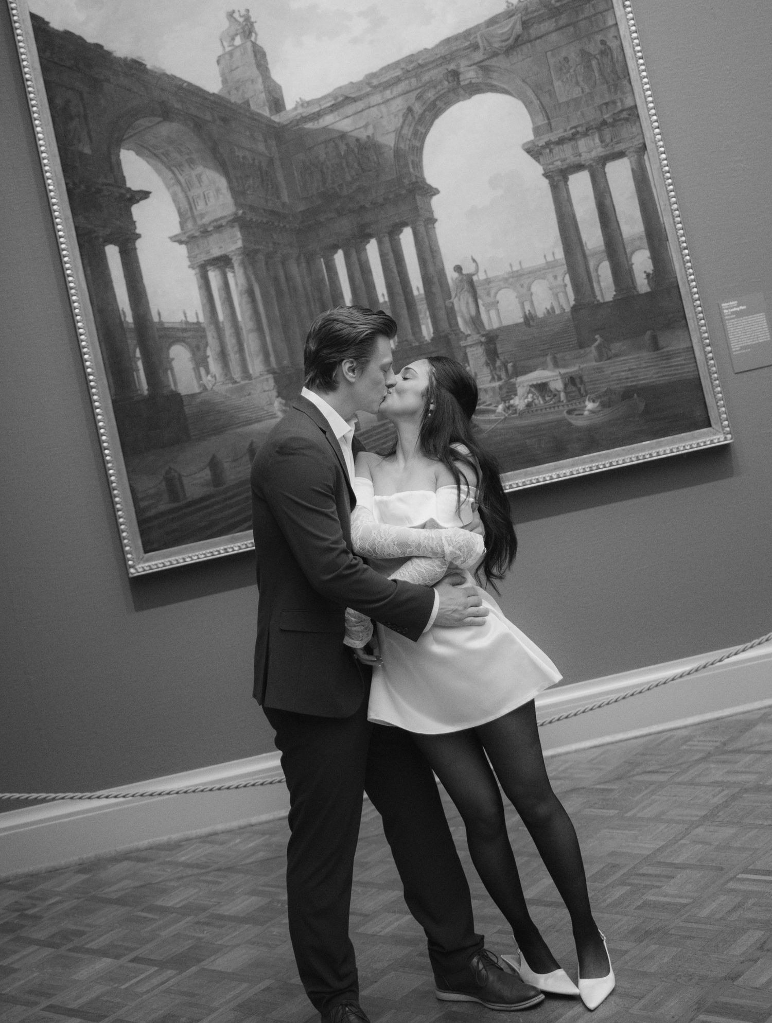 A couple embraces in front of a black and white classical painting in an art gallery.