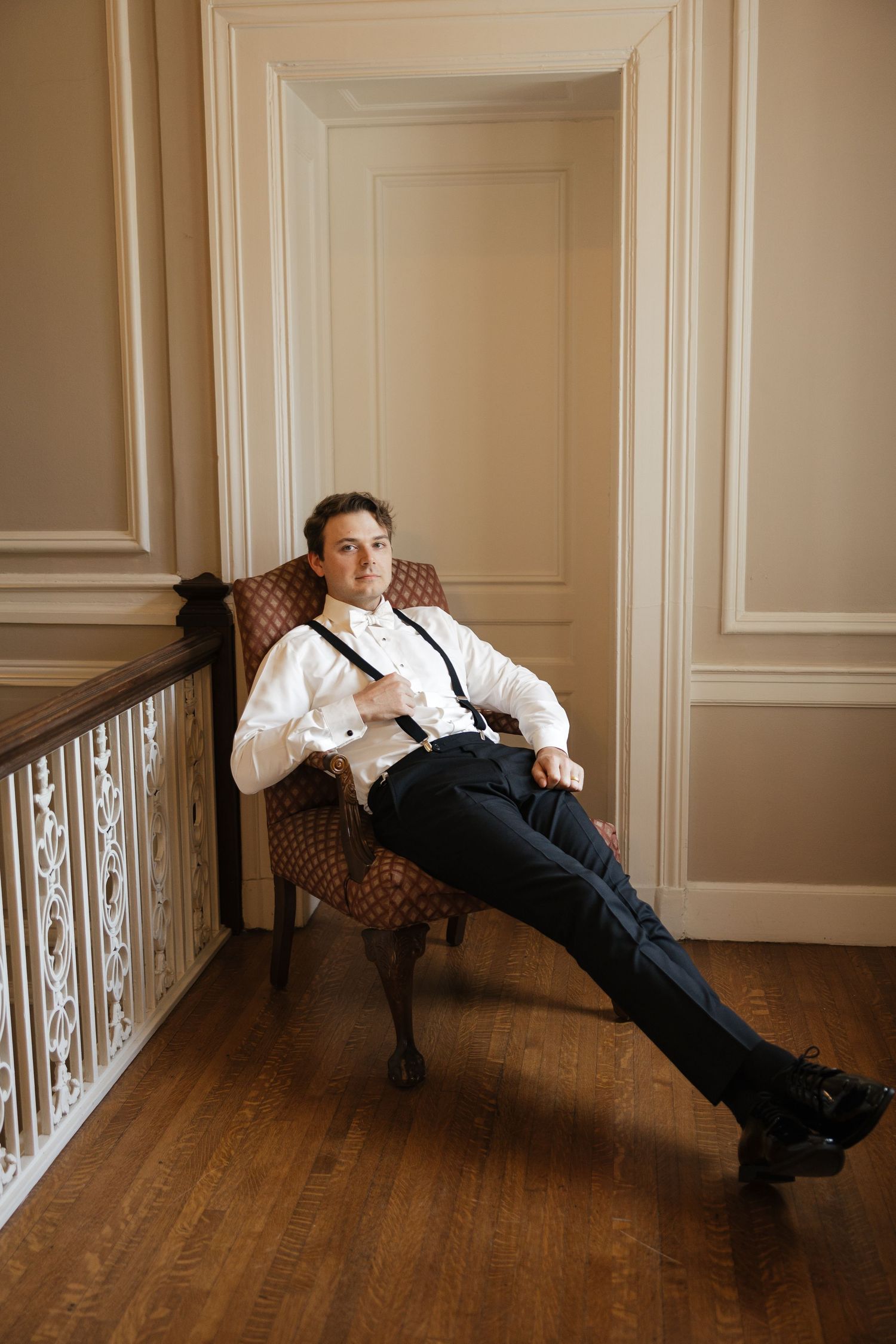 A dapper man in white shirt and suspenders relaxes in an ornate chair in an elegant room with wood floors.