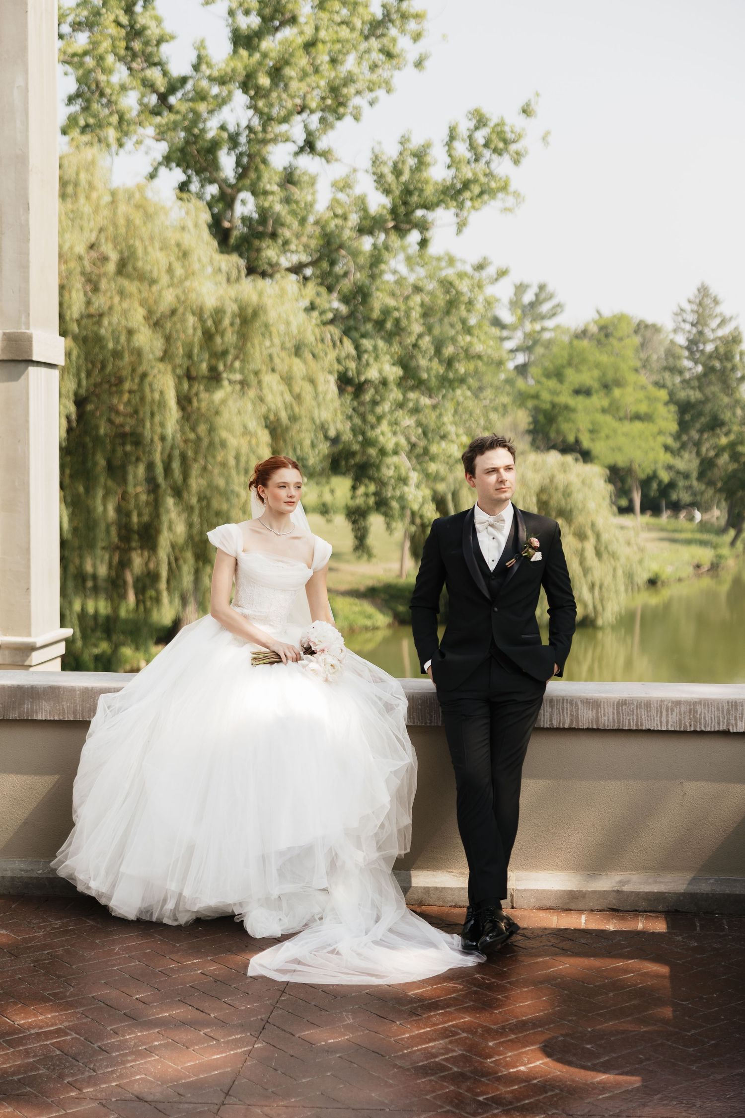 Bride and groom pose by stone balustrade overlooking serene pond with weeping willow at wedding venue.