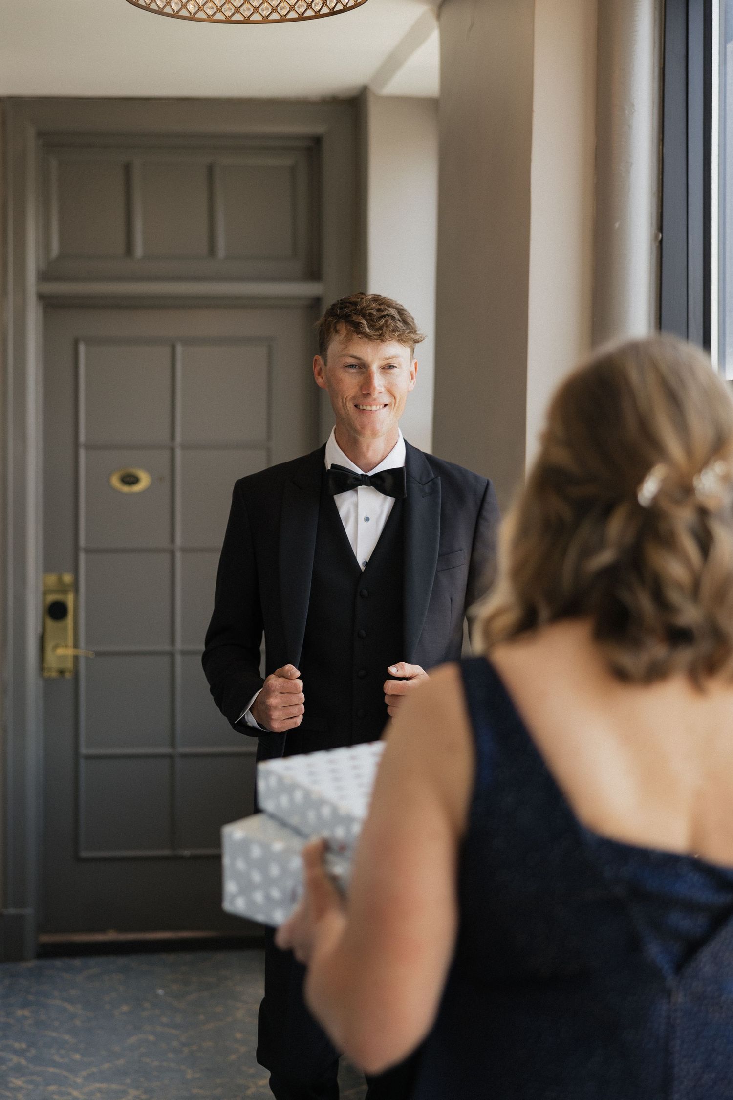 Young man in black tuxedo and bow tie smiles at woman in navy dress holding a gift box at a formal event.
